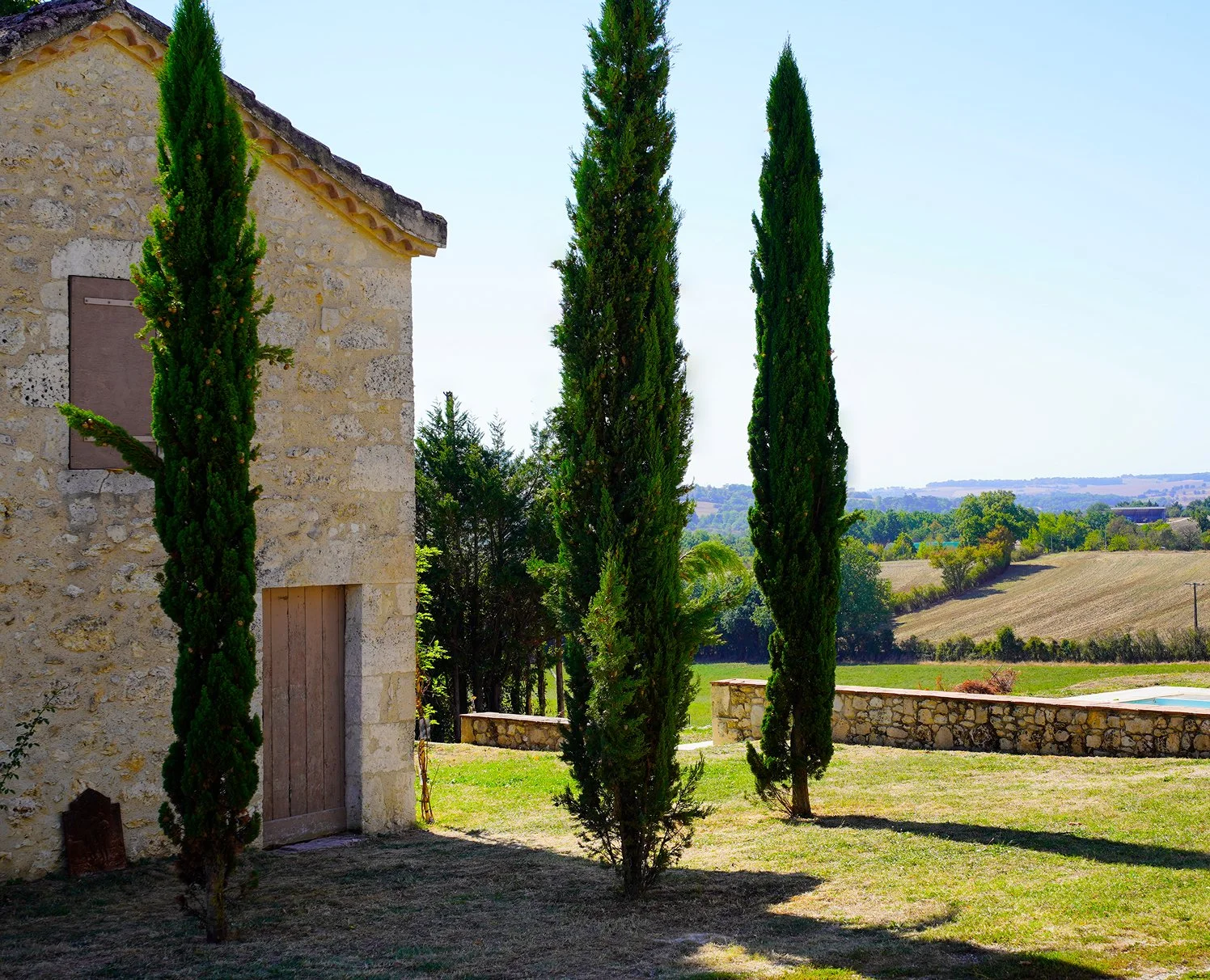 A stone house with a wooden door surrounded by tall, narrow cypress trees in a countryside landscape under a clear blue sky.