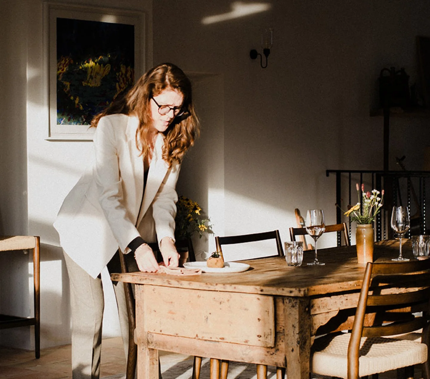 A woman in a cream-colored suit standing at a dining table, slicing a piece of meat or fish on a plate. The table is set with plates, wine glasses, and a small vase with flowers. Sunlight streams into the room, creating bright and shadowed areas.