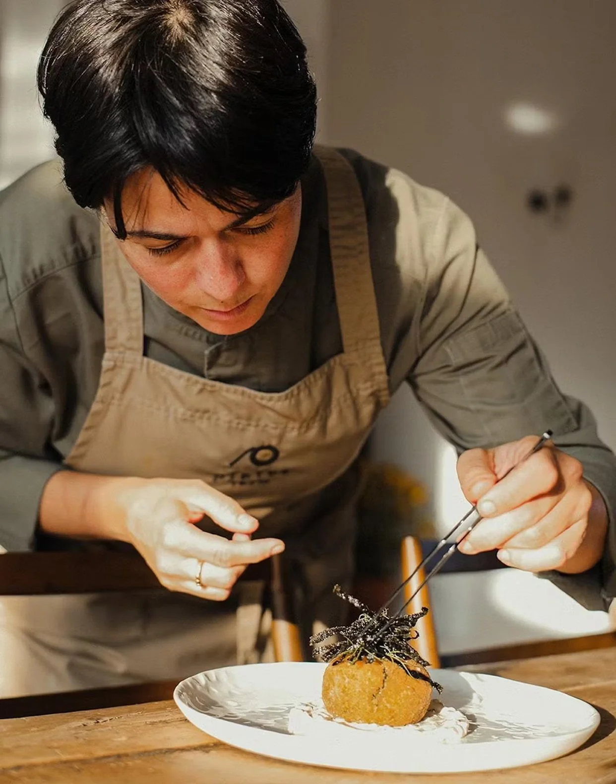 Chef garnishing a plated dish with tweezers, focusing on a crispy, golden-brown item topped with black garnish on a white plate.