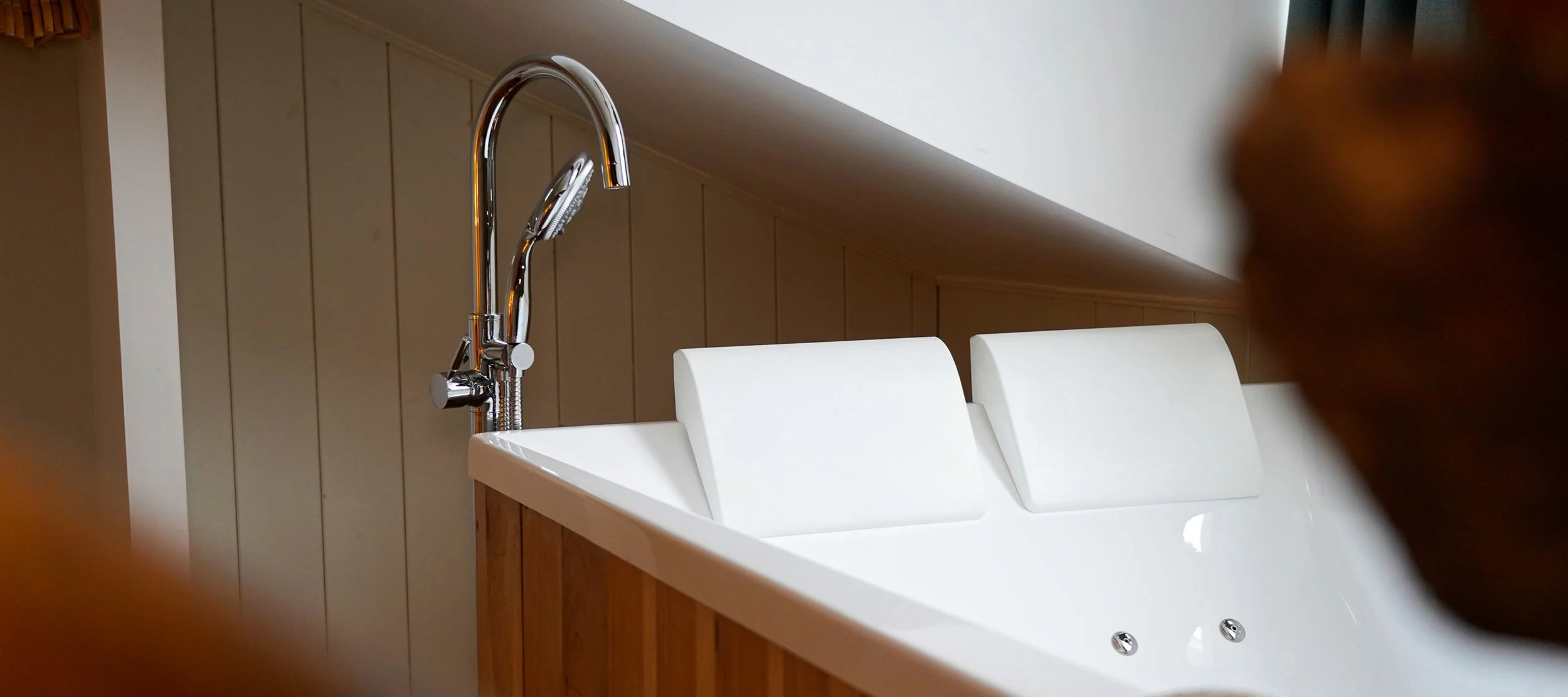 A close-up of a bathroom bathtub with a chrome faucet and a handheld showerhead, with a beige wooden panel behind and a white headrest on the tub.