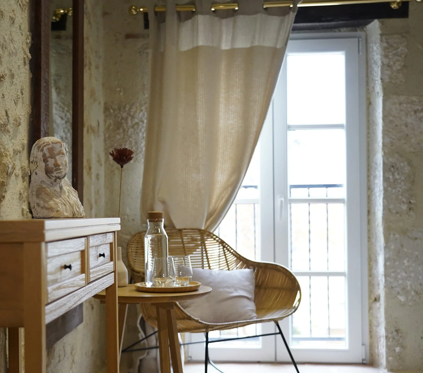 A cozy interior corner with a wooden side table, a glass water pitcher, and a wicker chair with a white cushion against a stone wall, next to a large window with beige curtains.