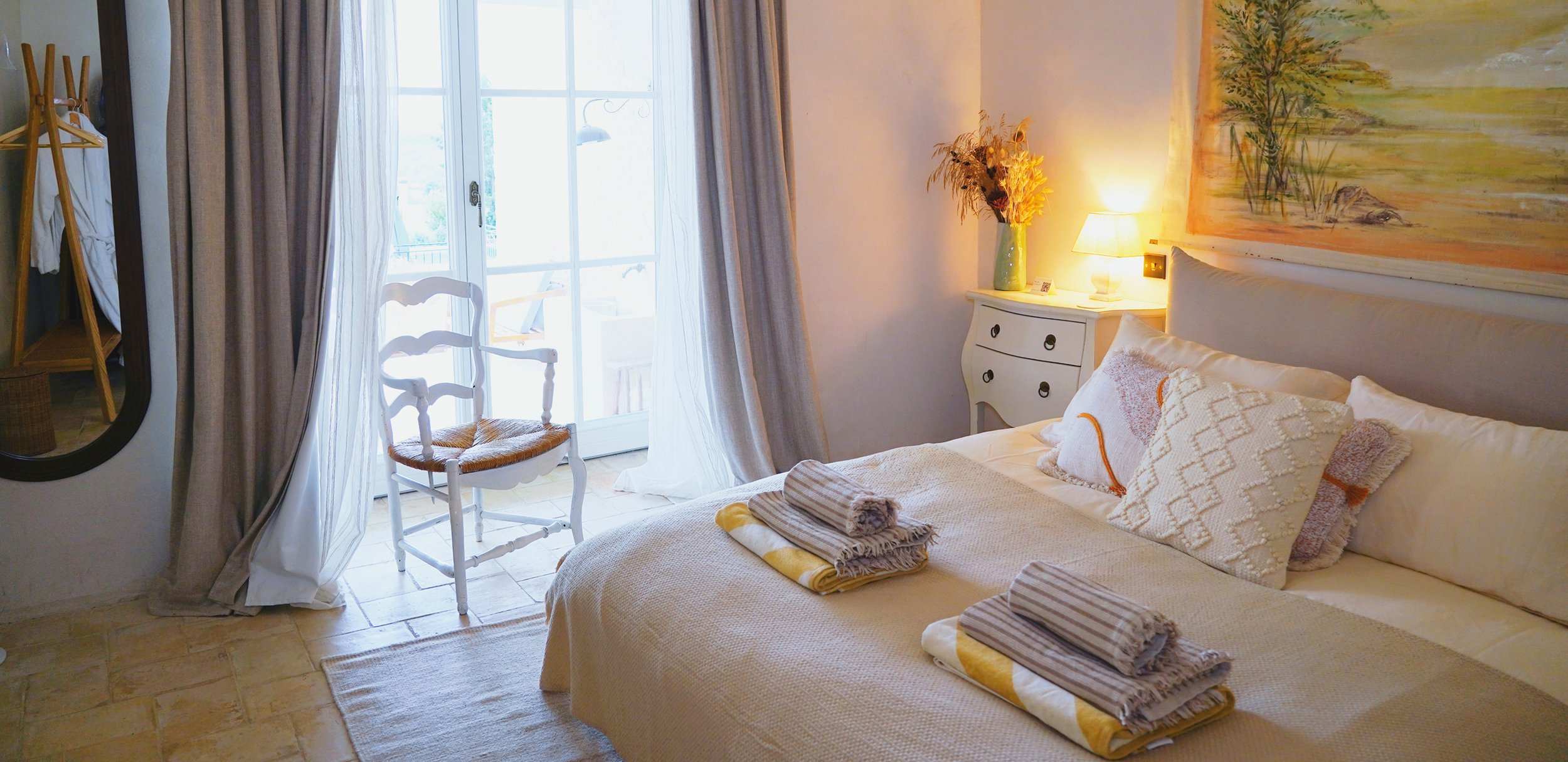 A cozy bedroom with a bed decorated with plush pillows and folded towels, a white dresser with a vase of dried flowers, a yellow-lit table lamp, and a large window with curtains overlooking a balcony.