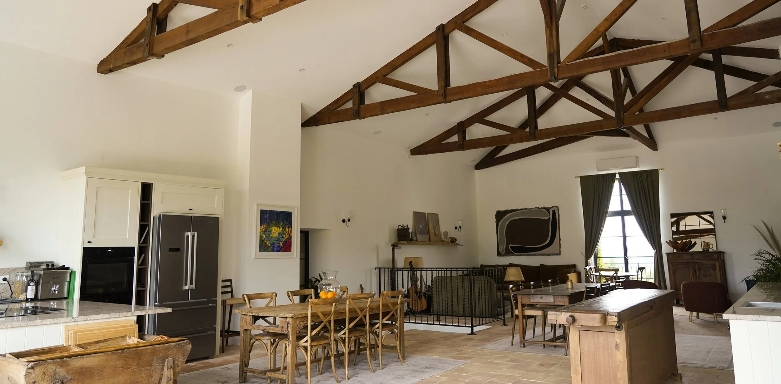 Open-concept living and dining room with exposed wooden beams, a large window with green curtains, and various furniture including a wooden dining table, green sofa, armchair, and sideboard.