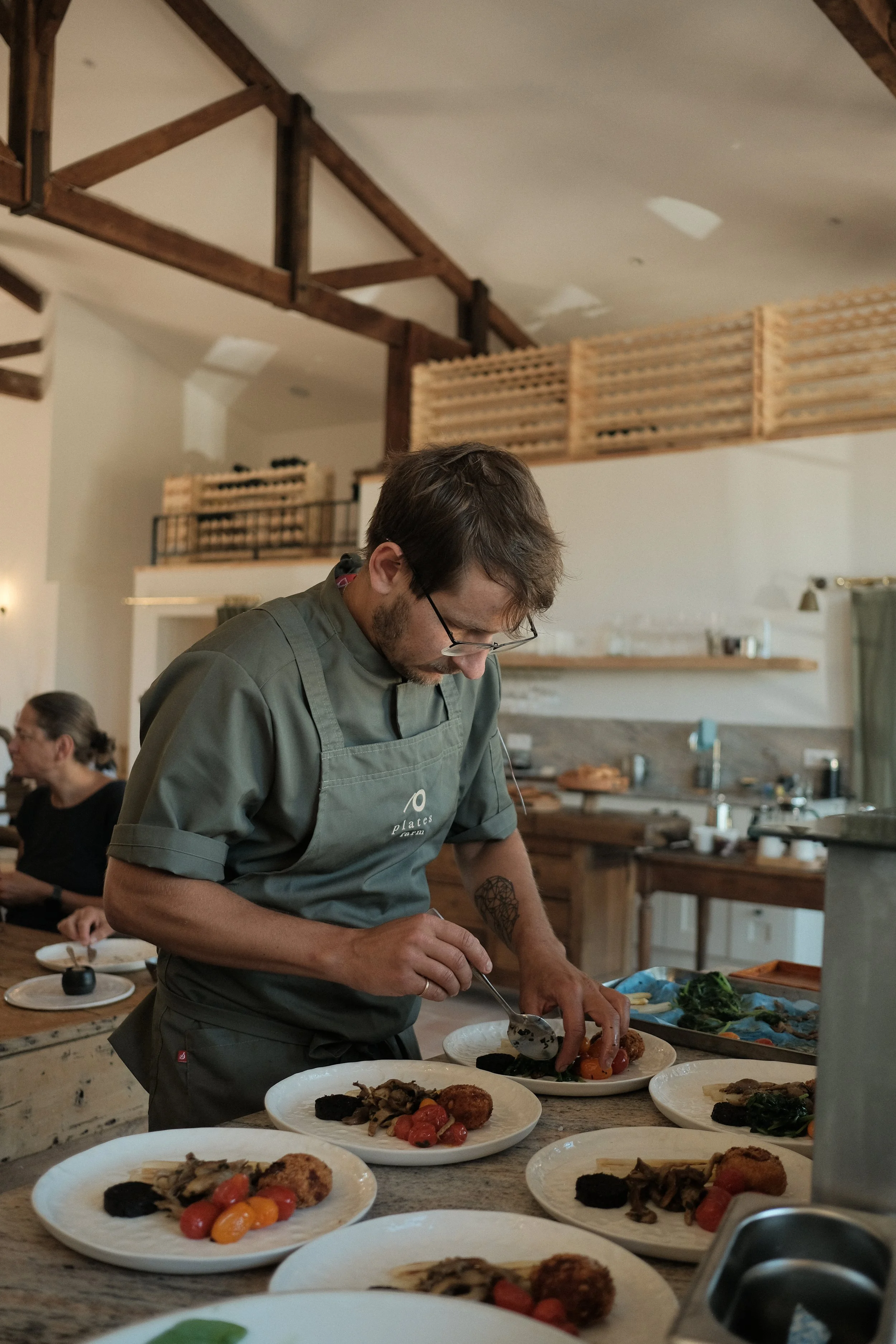 A chef in a gray apron is plating food in a rustic kitchen with wooden beams and a loft. Plates of food with cherry tomatoes, mushrooms, and fried items are arranged on the counter around him.