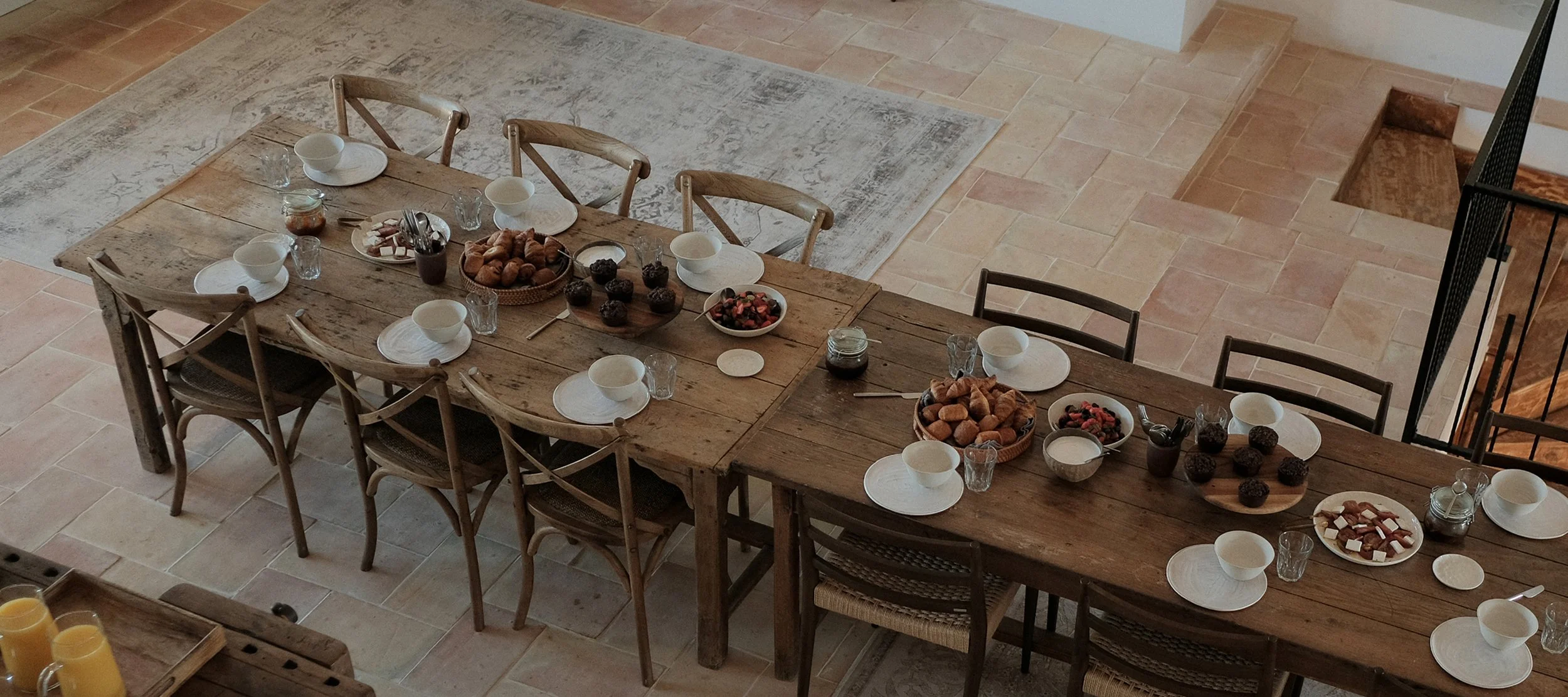 A long wooden dining table set for breakfast with plates, bowls, glasses, and cutlery, decorated with bowls of muffins, berries, and honey jars, in a room with tiled floors and a rug underneath part of the table.