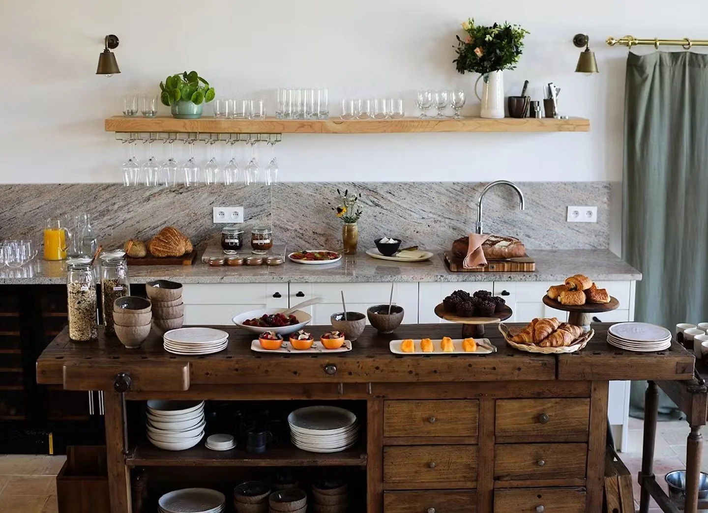A kitchen with a rustic wooden island, various baked goods, fruits, and breakfast items laid out on the counter, a marble backsplash with electrical outlets, white cabinets, a wall-mounted wooden shelf with glasses and plants, and a light-colored curtain to the right.
