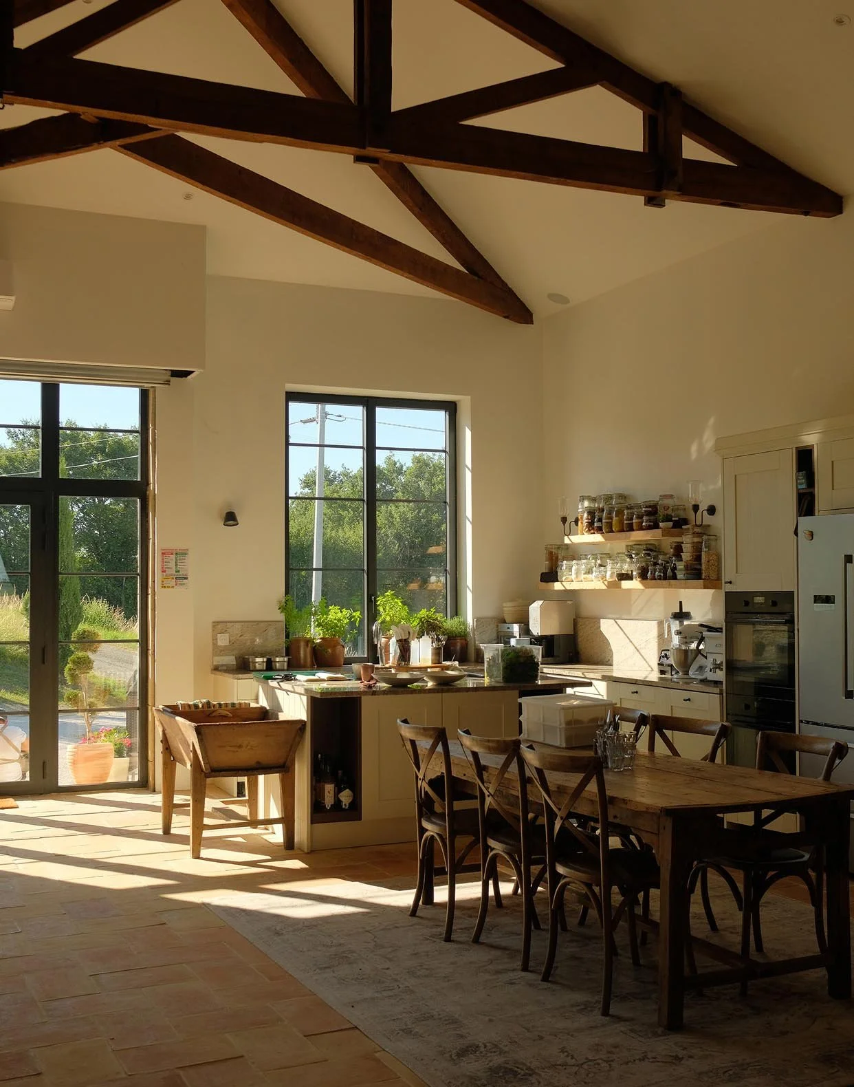 Sunlit kitchen with large windows, wooden table with chairs, cabinets, and open shelves with jars and plants, featuring exposed wooden beams.
