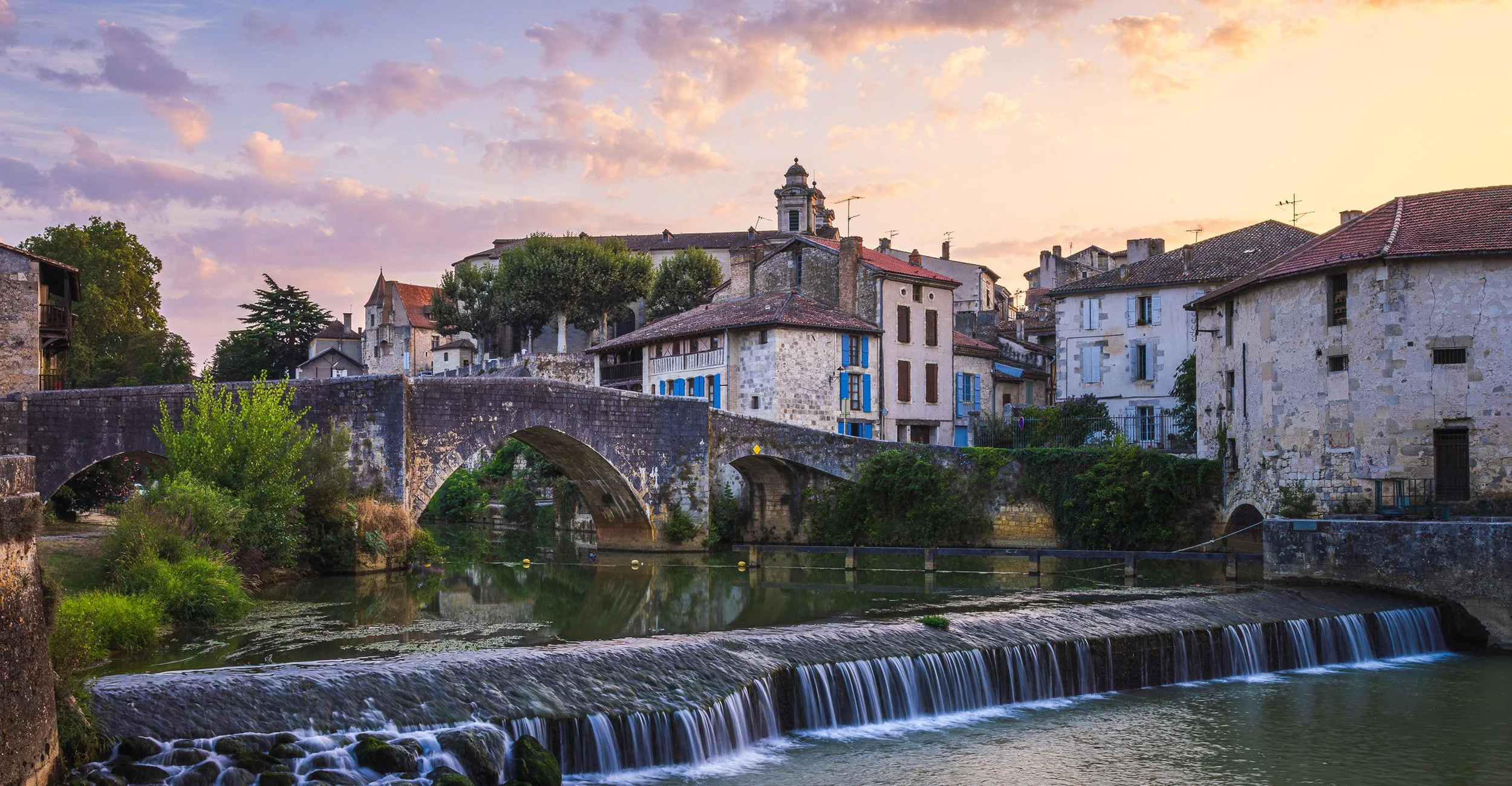 View of a small town with old stone buildings, a stone bridge over a river, and a waterfall at sunset.