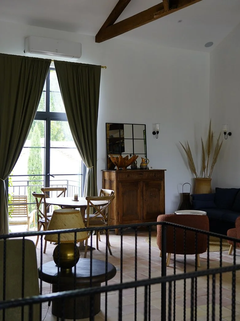 Living room with large window and green curtains, wooden dining table with chairs, vintage wooden cabinet, mirror, black sofa, armchair, dried pampas grass in a vase, and a small table, with natural light coming in.