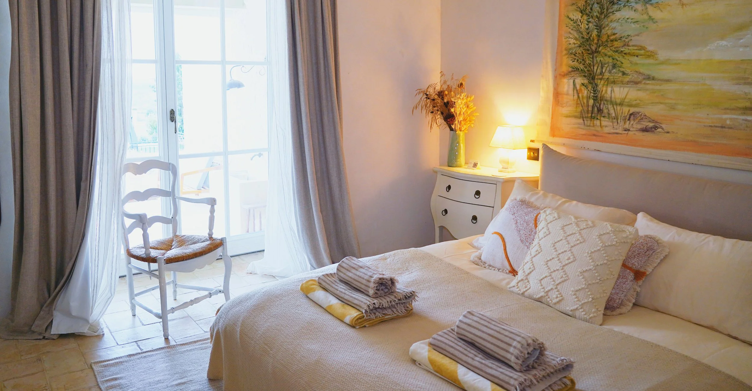 A cozy bedroom with a large bed, decorative pillows, and neatly folded towels, near a window with curtains, a white chair, a nightstand with a vase of flowers, a lamp, and a colorful painting on the wall.