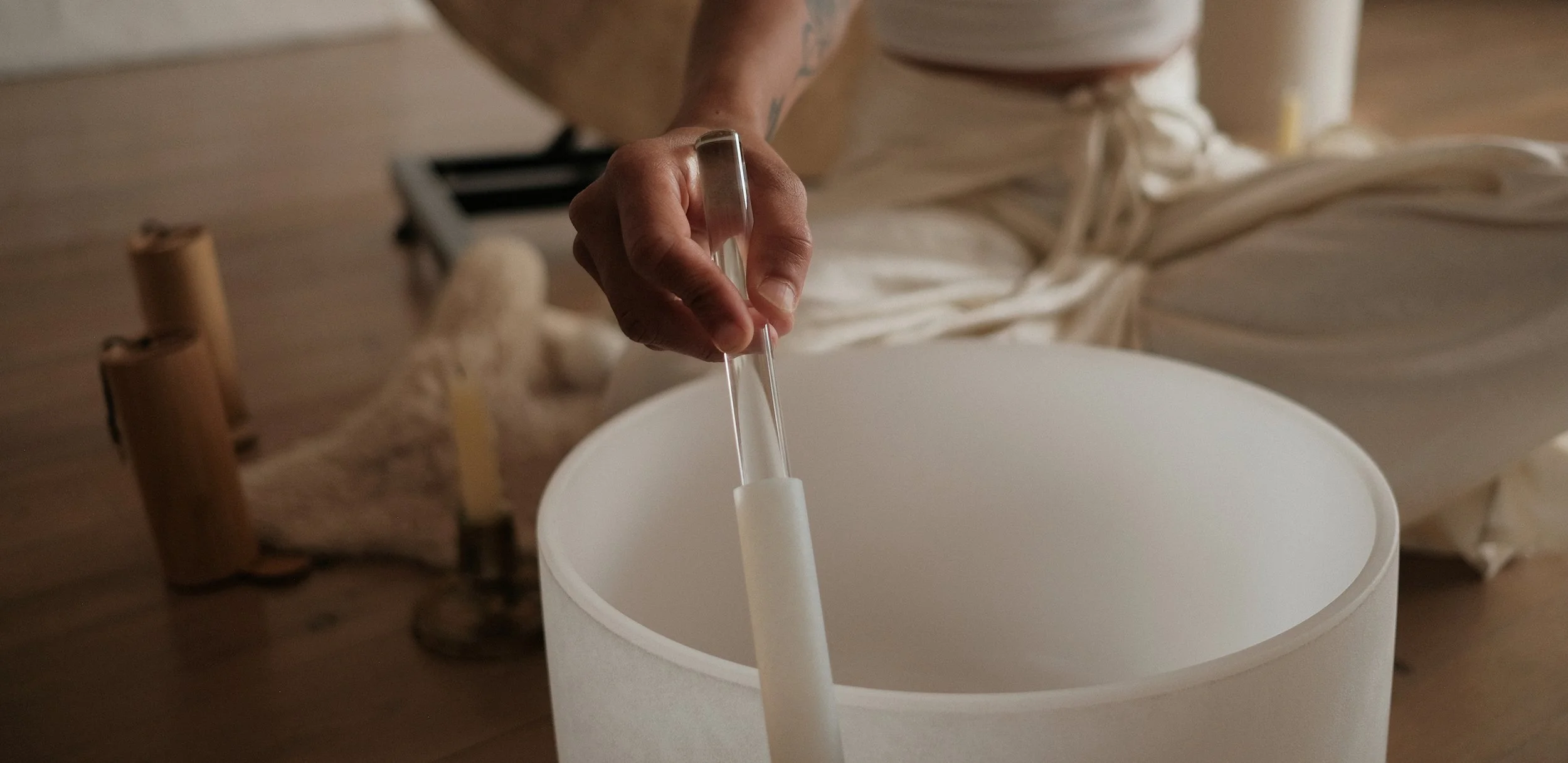 A person is playing a white acoustic drum or cymbal with a drumstick. They are kneeling on the wooden floor, and their torso, including tattoos on their arm, is visible. The person is wearing cream-colored pants tied at the waist, and there are various percussion instruments visible in the background.