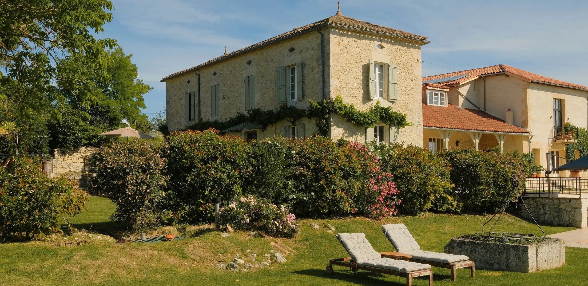 A large stone house with shutters, surrounded by greenery and colorful bushes, with two lounge chairs and an umbrella in the backyard on a sunny day.