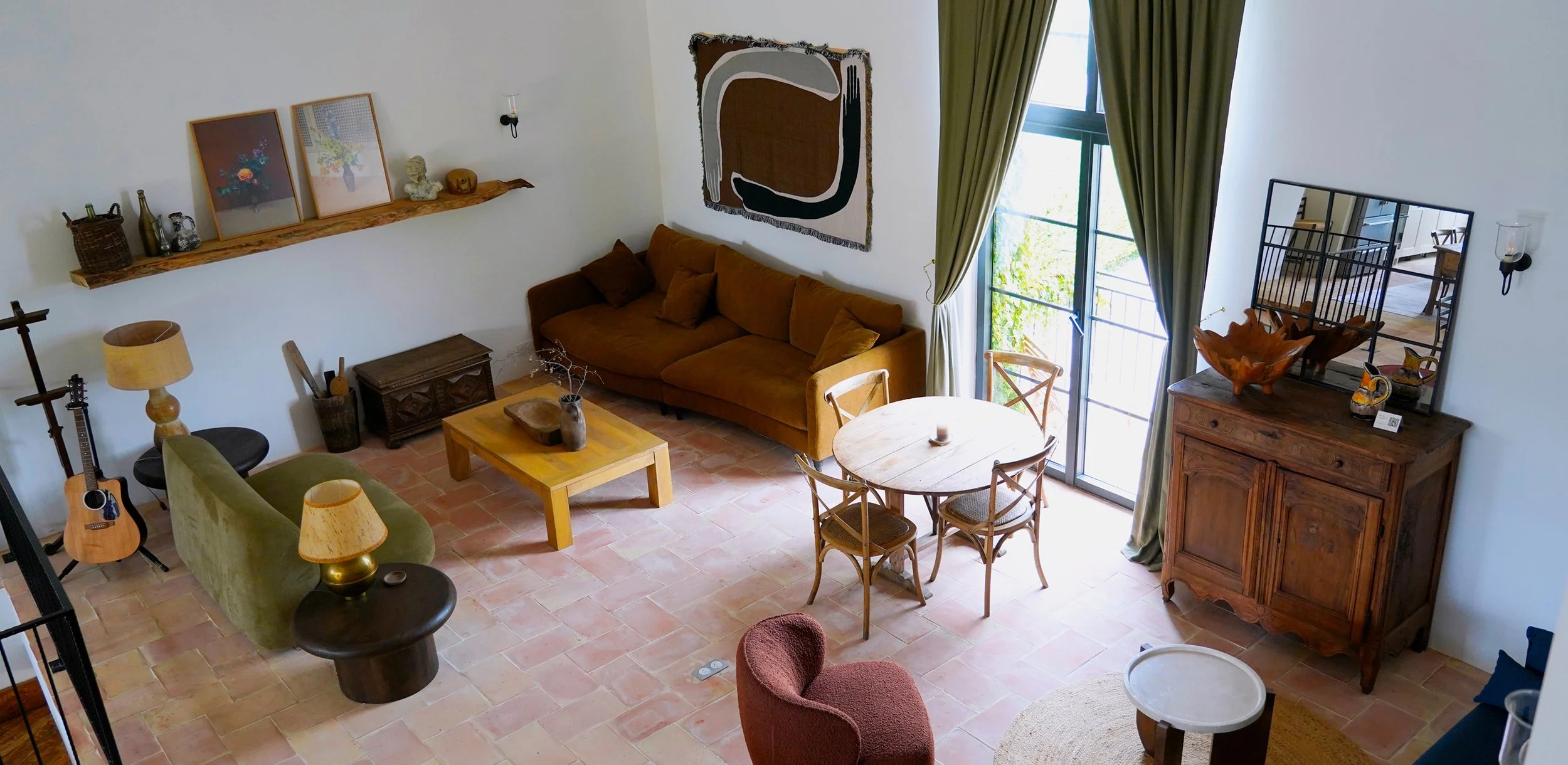 Living room with brown sofa, round dining table with chairs, wooden cabinet with mirror, large window with green curtains, hanging artwork, and various decorative items.