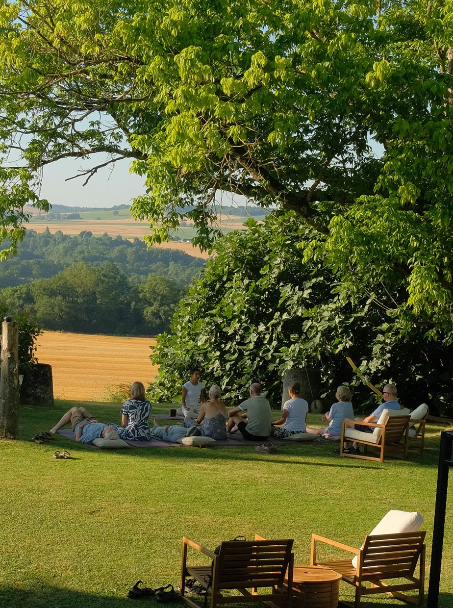 People gathered outdoors under a large tree, seated on blankets and chairs, engaging in a group activity with a scenic countryside background.