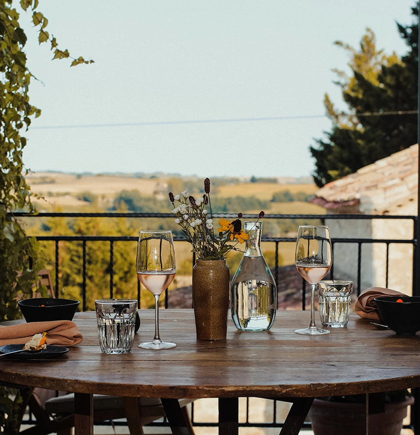 A wooden outdoor dining table set with three glasses of rosé wine, two clear water glasses, black bowls, orange napkins, and a brown vase with wildflowers. A glass pitcher of water is also on the table, with a scenic view of rolling hills and trees in the background.