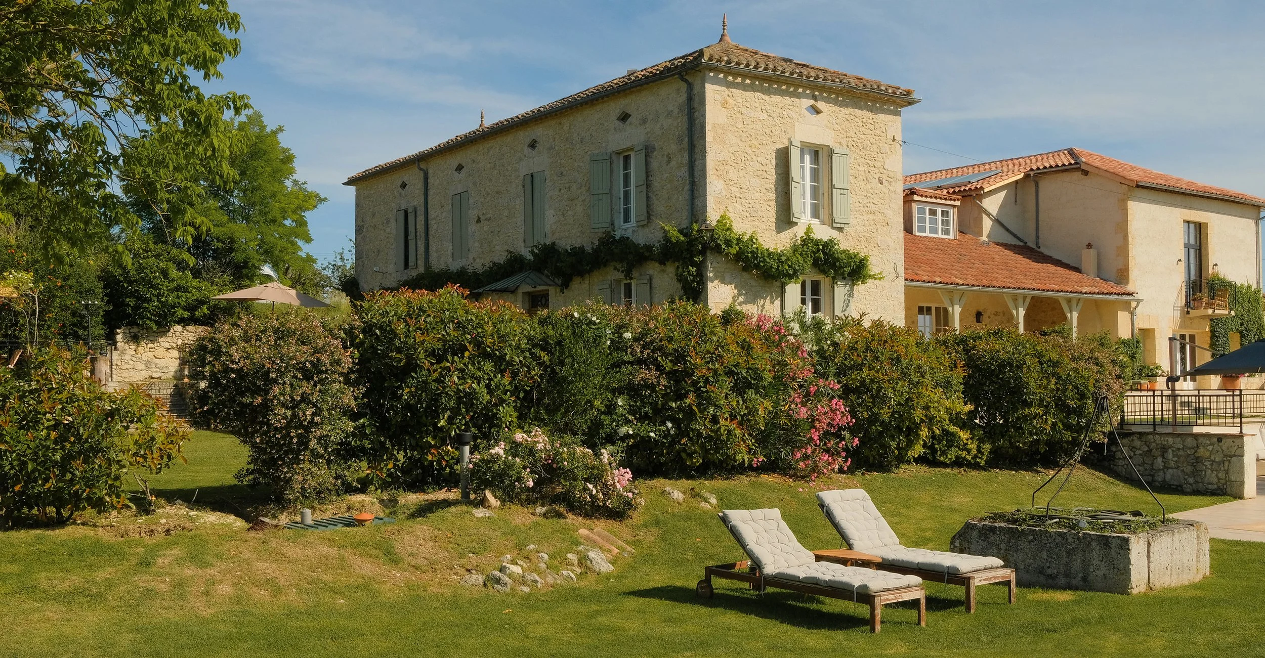 A large stone house with green shutters and a tiled roof, surrounded by lush greenery, flowering bushes, and two lounge chairs on a well-manicured lawn.