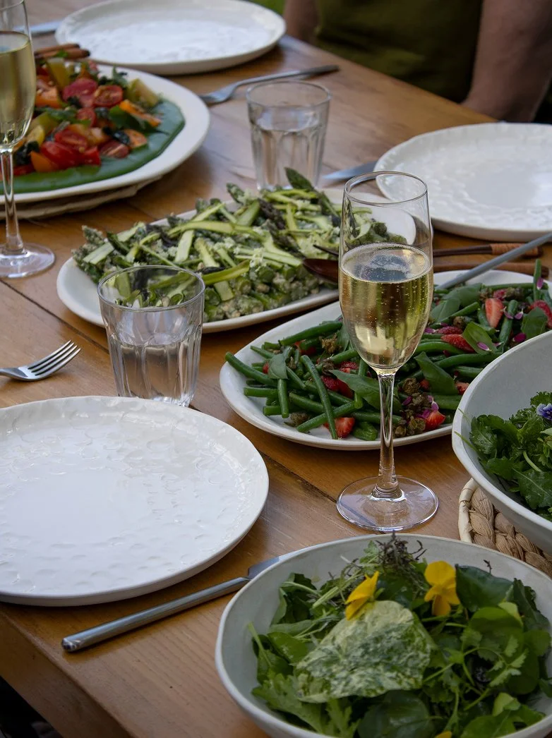 A table set with empty white plates, several dishes of salad with vegetables, and glasses of sparkling wine. There are two glasses of wine, one nearly full, and glasses of water. The salads include mixed greens, green beans, and a zucchini salad.