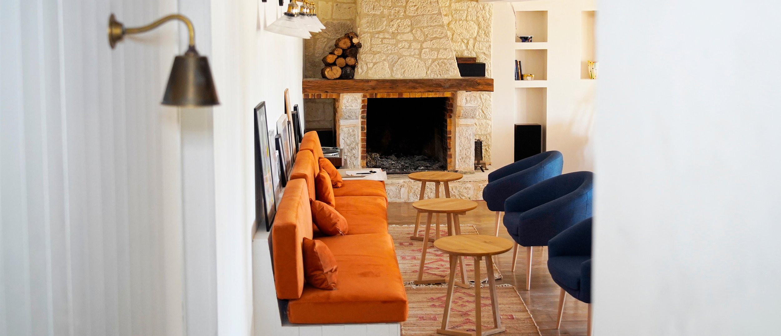 Living room view through doorway, featuring an orange sofa, blue armchairs, a stone fireplace with wood logs, wooden coffee tables, built-in shelves, and wall-mounted lights.