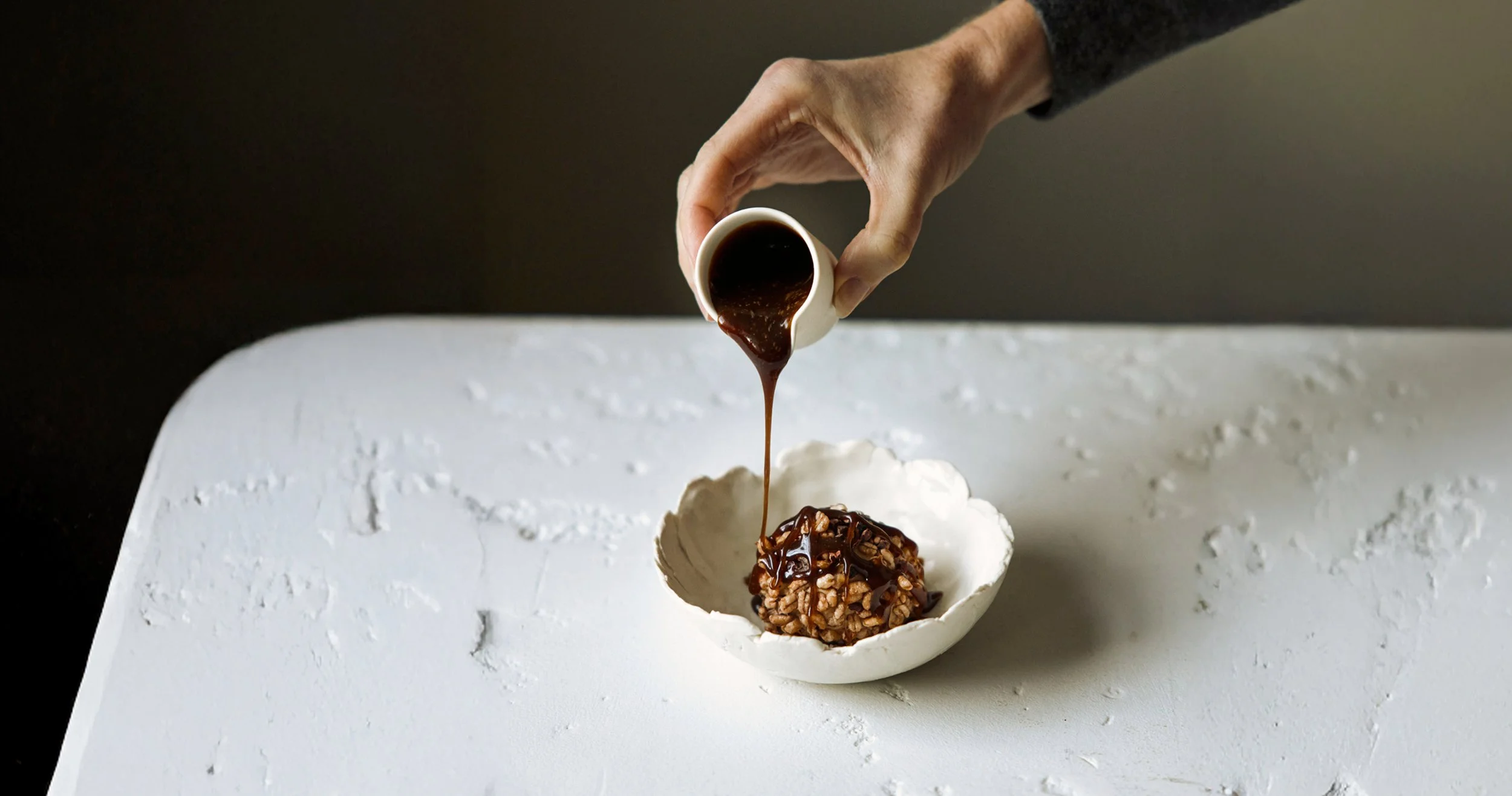 A hand pouring chocolate sauce over a rice cereal treat on a white plate.