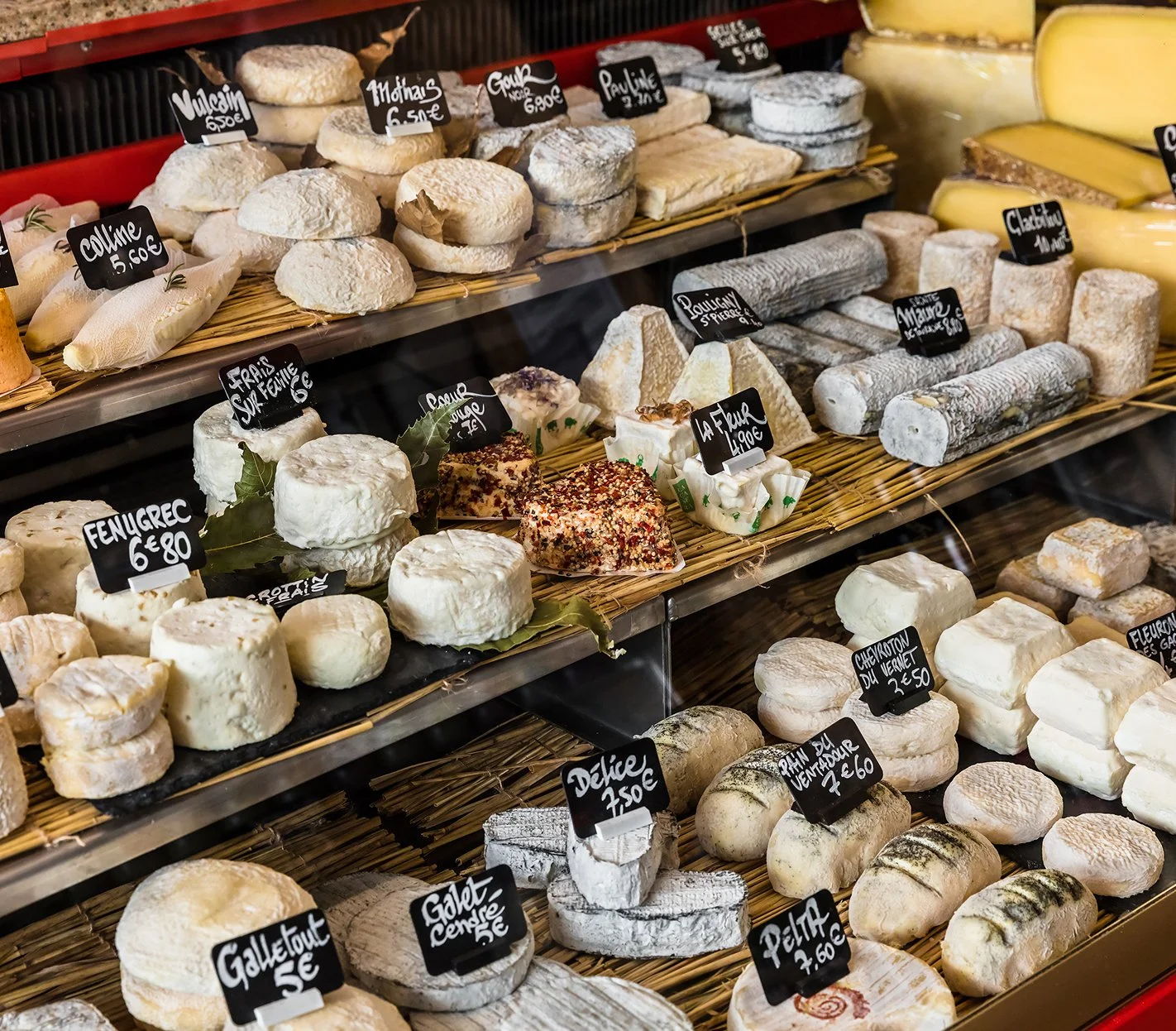 Display case with various cheeses on shelves, labeled with small black signs showing names and prices. The cheeses come in different shapes and sizes, including wheels, logs, and blocks.
