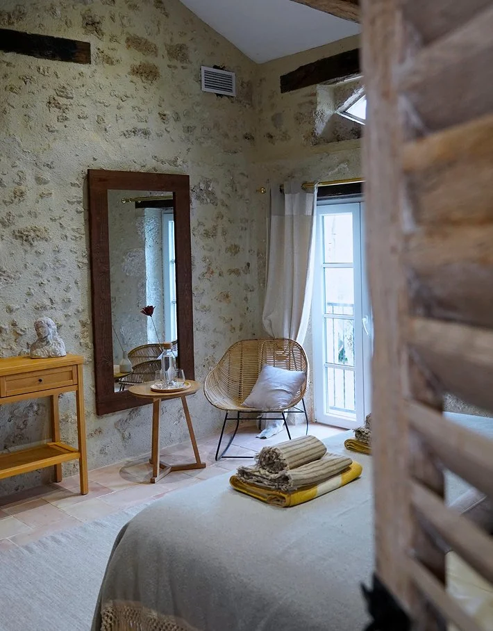 Cozy bedroom with stone walls, a bed with folded towels on top, a wicker chair by a glass door with curtains, a wooden side table with a vase and decorative items, and a large mirror on the wall.
