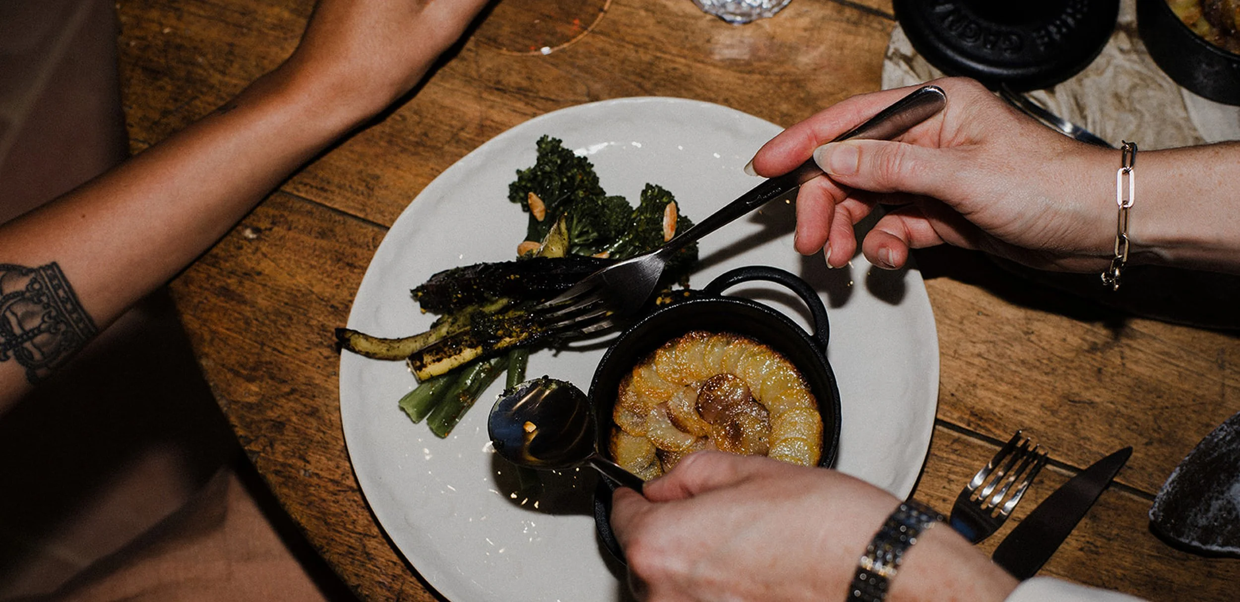 Person using a fork to eat grilled fish and vegetables on a white plate at a wooden table