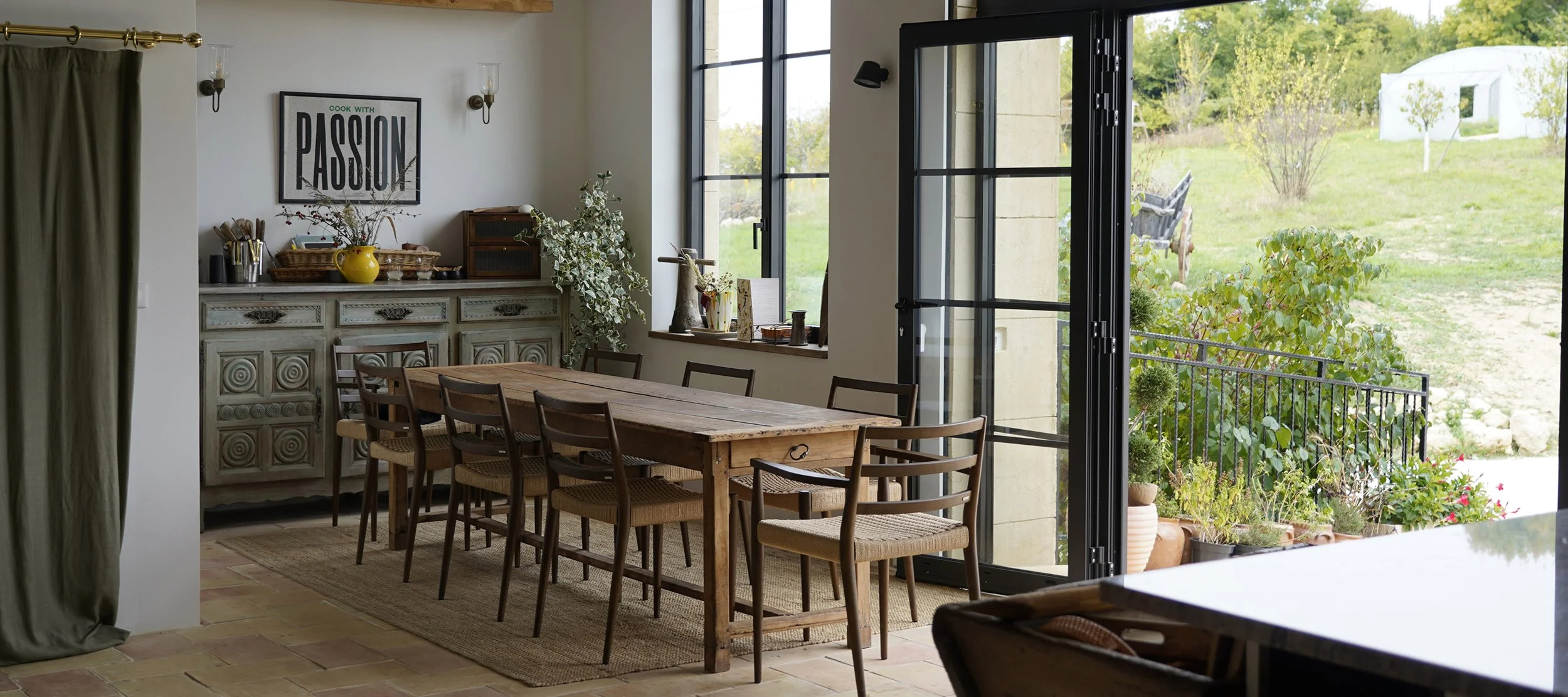 Dining room with wooden table and chairs, large open glass doors leading to a green outdoor area, decorative sideboard, framed artwork, and potted plants.