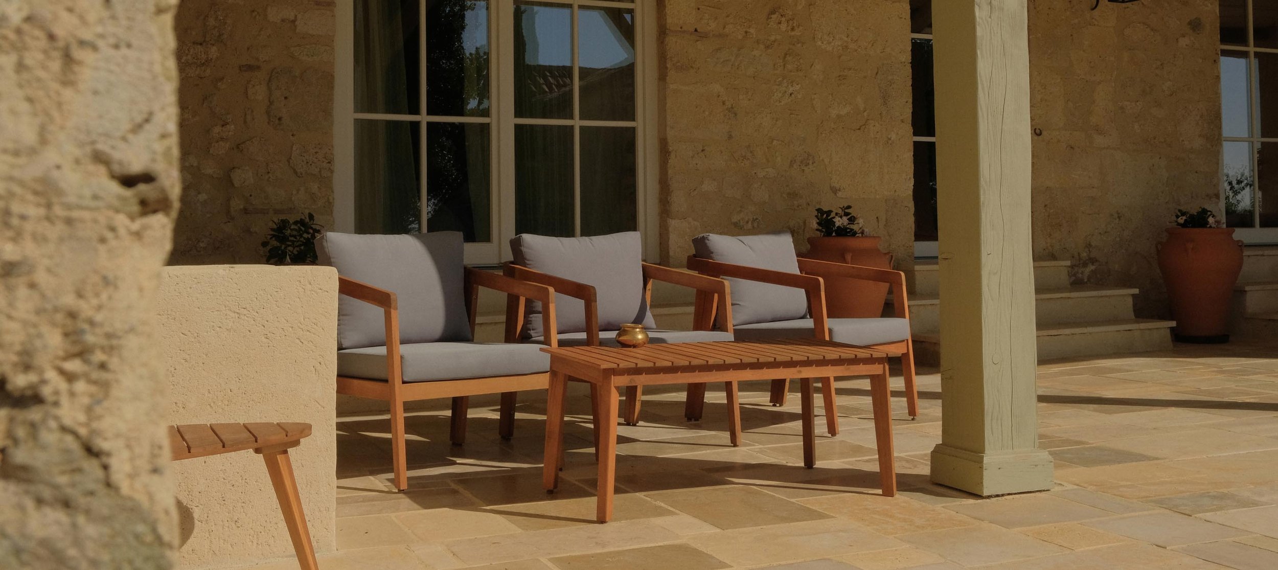 Outdoor patio area with wooden chairs, a small wooden table, potted plants, and stone steps leading up to a door with large windows.