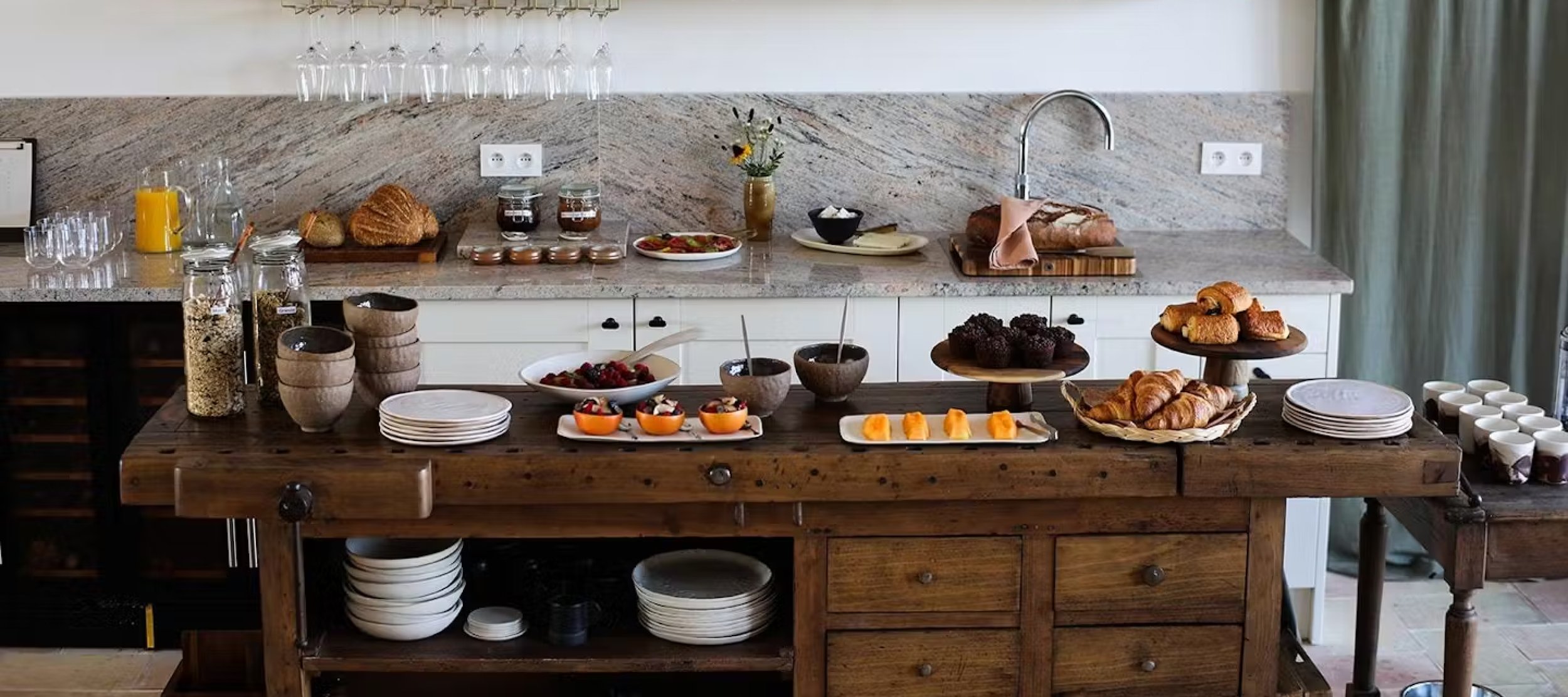 An assortment of breakfast items on a kitchen island and counter, including croissants, muffins, berries, yogurt, sliced cheese, bread, and jars of jam, honey, and oats.