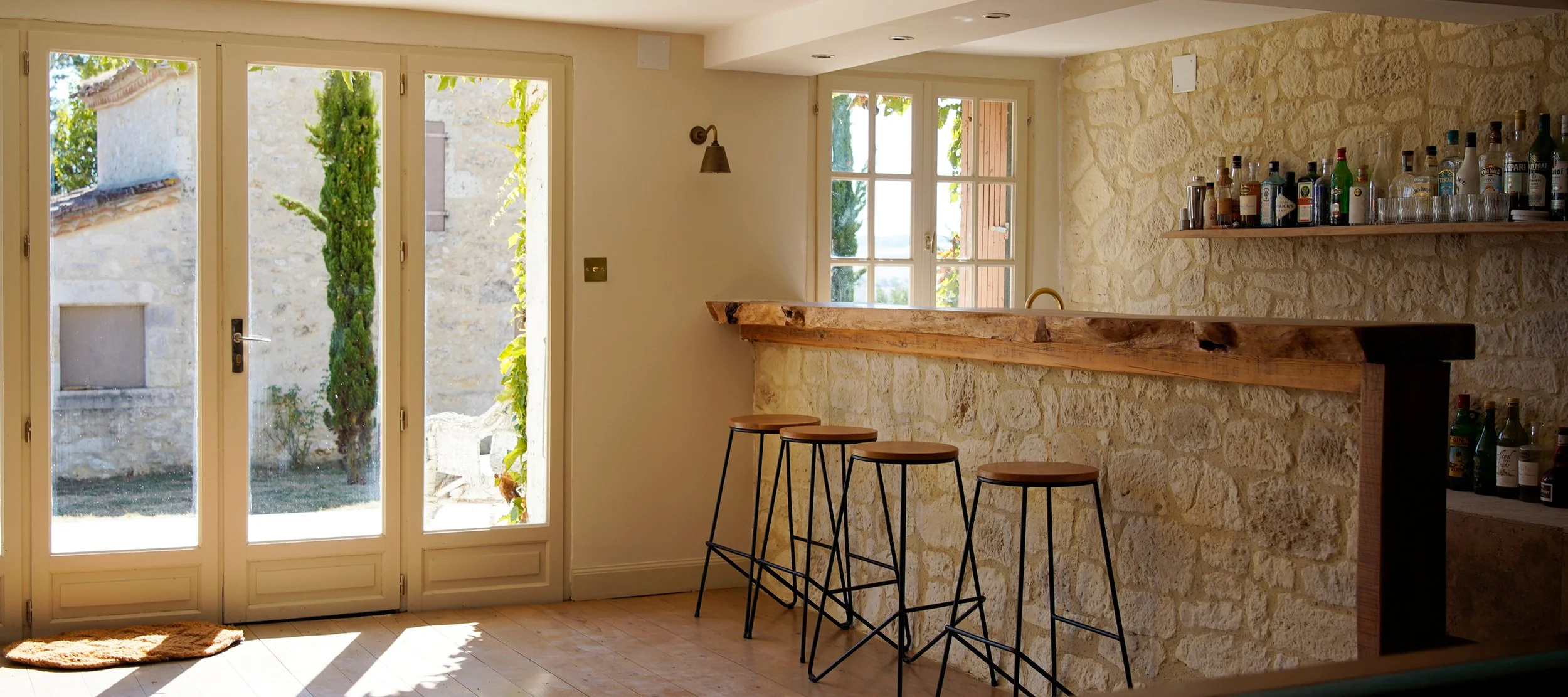 Interior of a cozy home bar area with a stone wall, wooden bar counter, and three bar stools. Large glass doors and windows let in natural light, revealing a stone building and greenery outside.