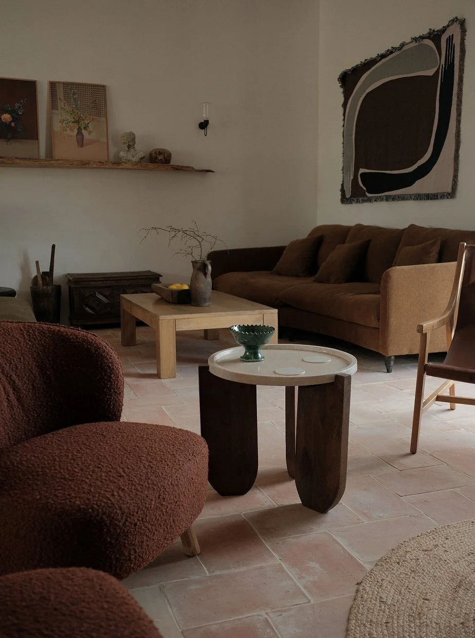 Living room with brown sofa, armchair, and two coffee tables, one with a green bowl and the other with a vase and decorative branch, artwork on the wall, terracotta tile floor, and neutral-colored walls.
