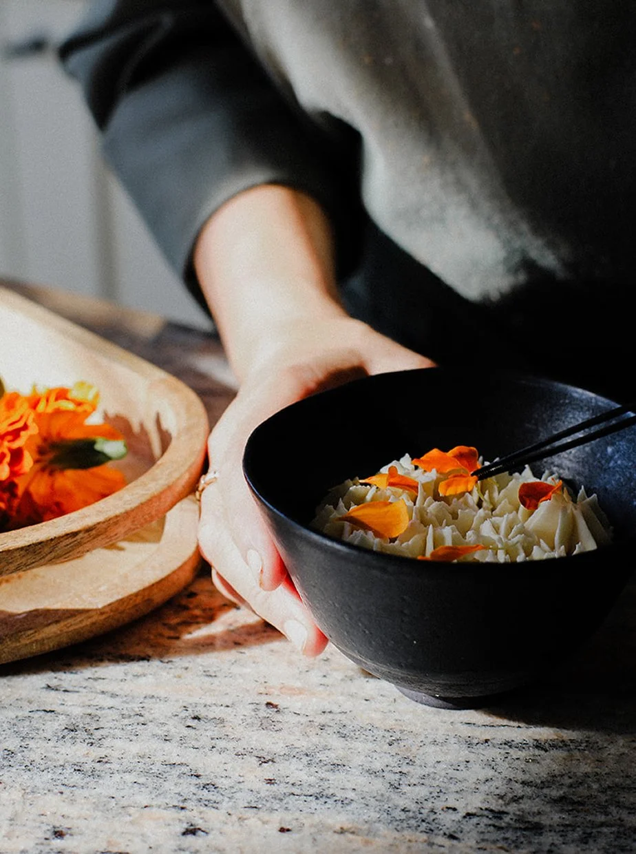 A person holding a black bowl filled with white pasta and orange flower petals. There is a wooden tray with orange flowers to the left on a speckled gray countertop.
