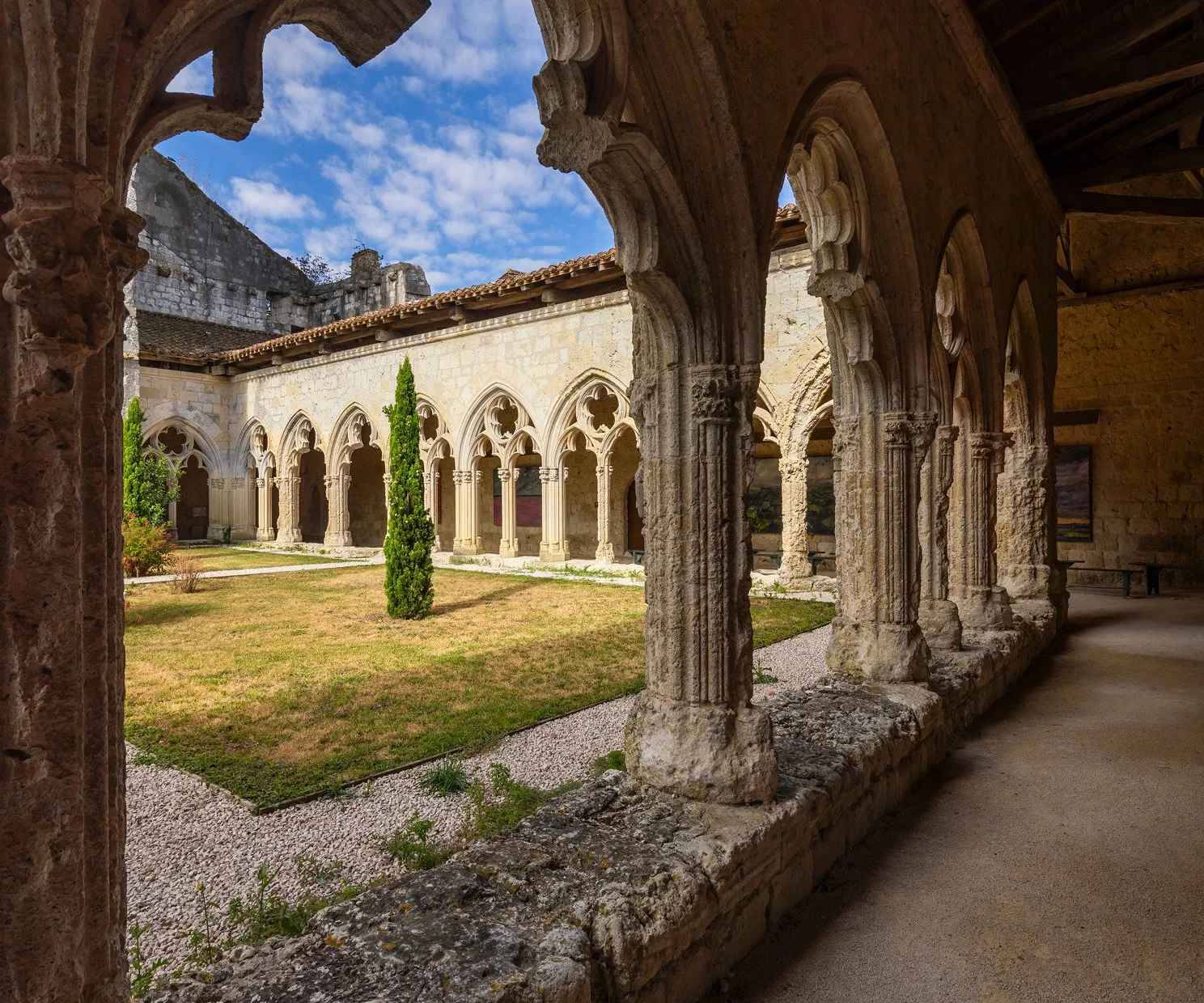 Ancient stone cloister with Gothic arches overlooking a courtyard with grass, a small cypress tree, and a stone wall with a tower in the background under a partly cloudy sky.