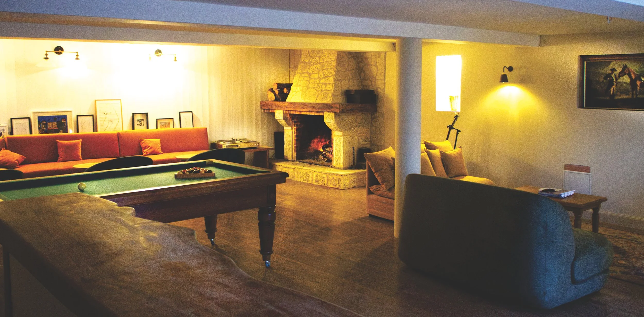 Cozy living room with a pool table, a fireplace, a long orange sofa with throw pillows, a vintage armchair, and framed artwork on the wall, illuminated by warm lighting.