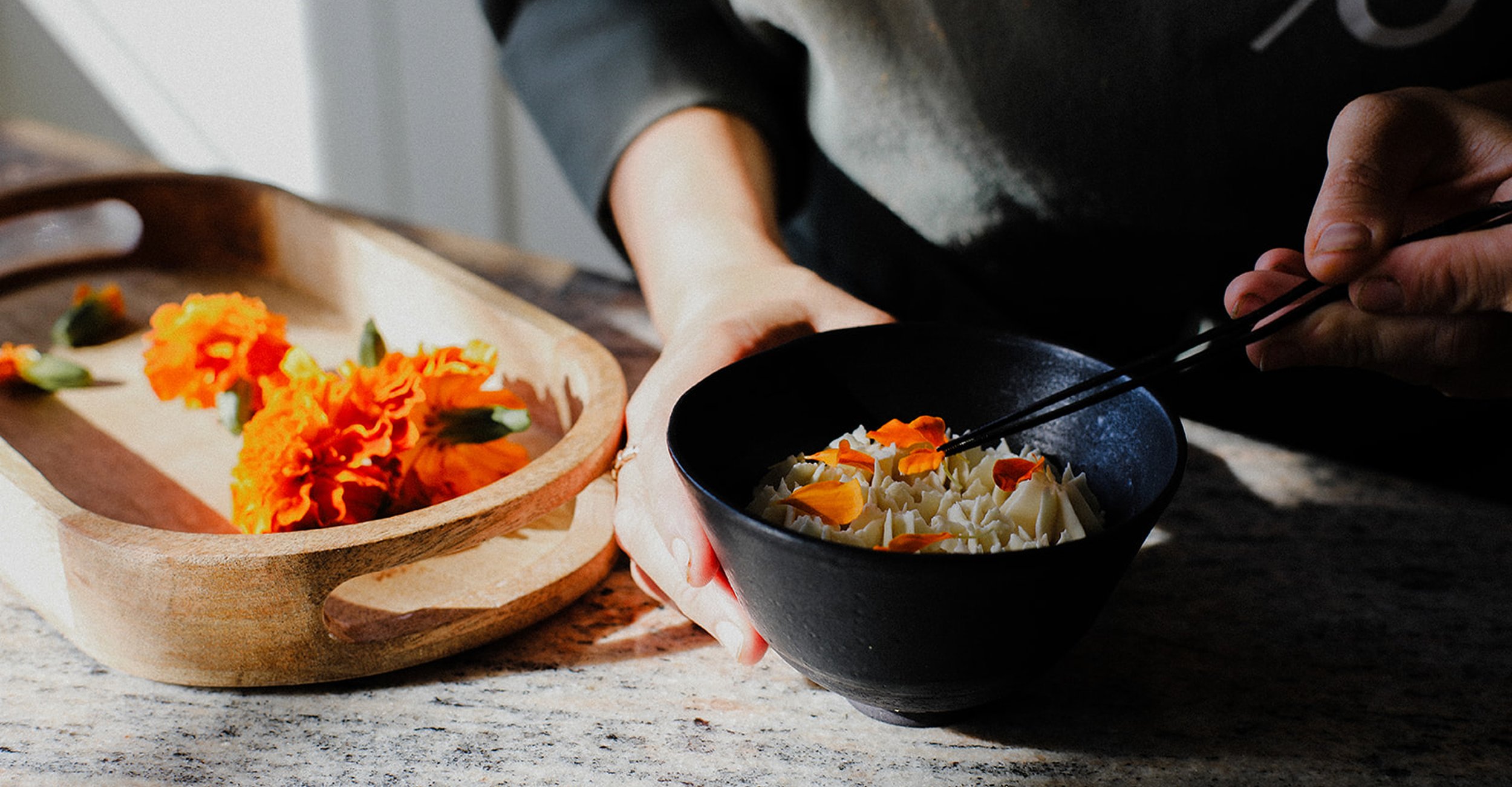 Person using chopsticks to serve a bowl of rice with flower petals. There is a wooden tray on the table with marigold flowers.