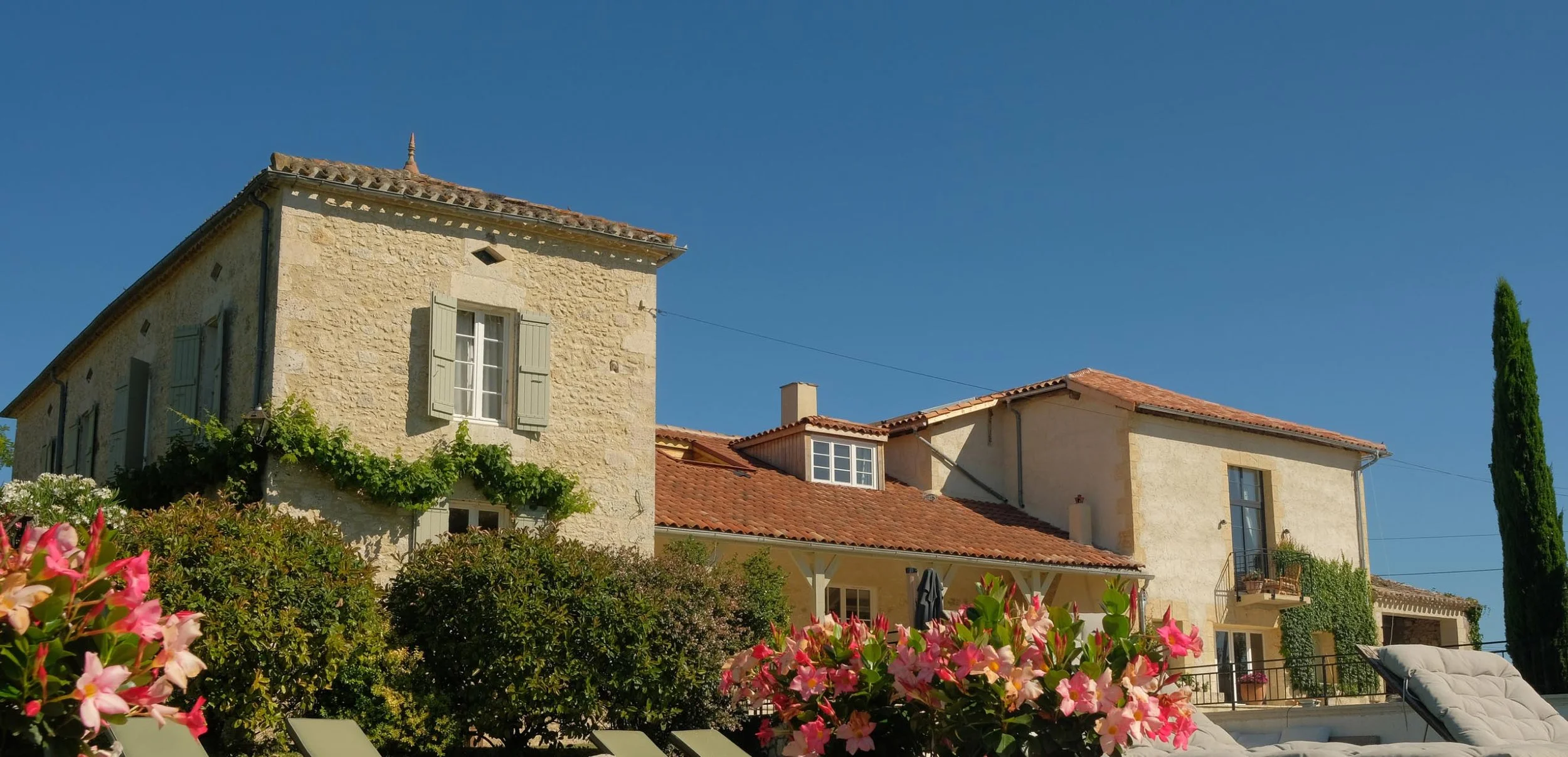 A Mediterranean-style house with light-colored stone walls, red-tiled roof, and multiple windows and balconies, surrounded by lush greenery and pink flowers under a clear blue sky.