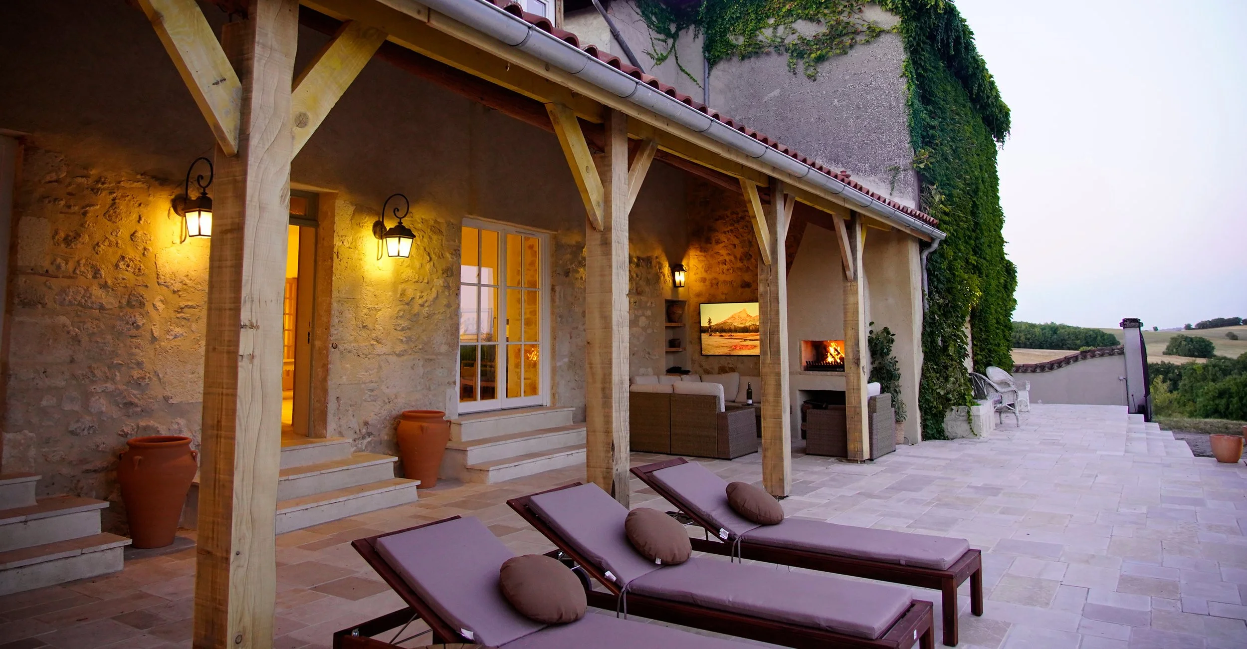 Outdoor patio area with lounge chairs, a fireplace with a fire burning, and a scenic view of rolling hills. The patio is attached to a rustic house with stone walls, wooden support beams, and outdoor lighting.