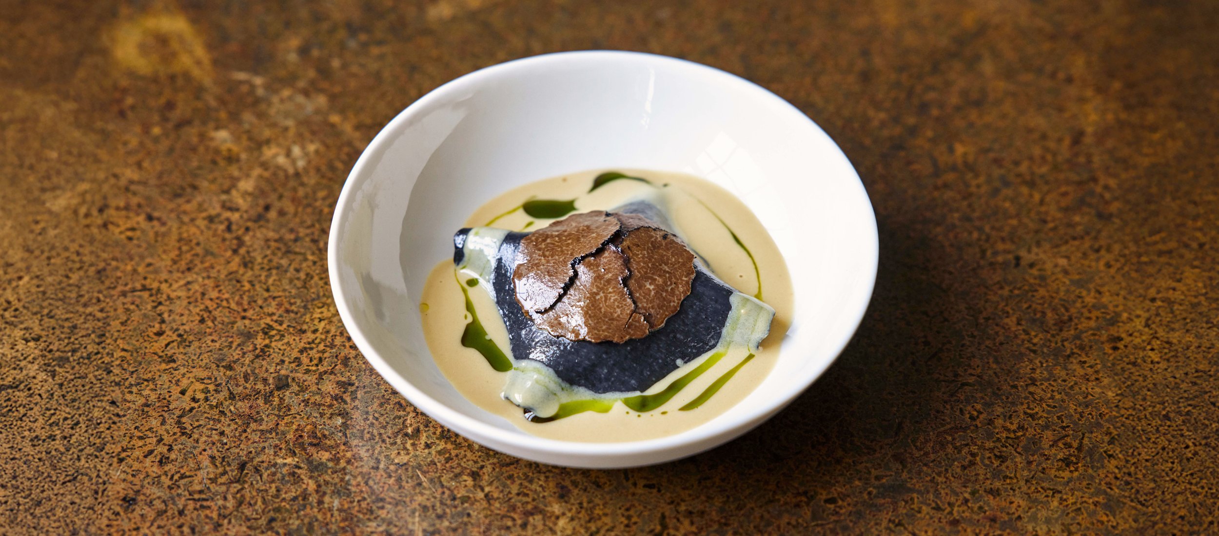 A dish featuring a piece of fish with a crispy brown skin, served in a creamy sauce with green oil drizzle in a white bowl on a textured brown surface.