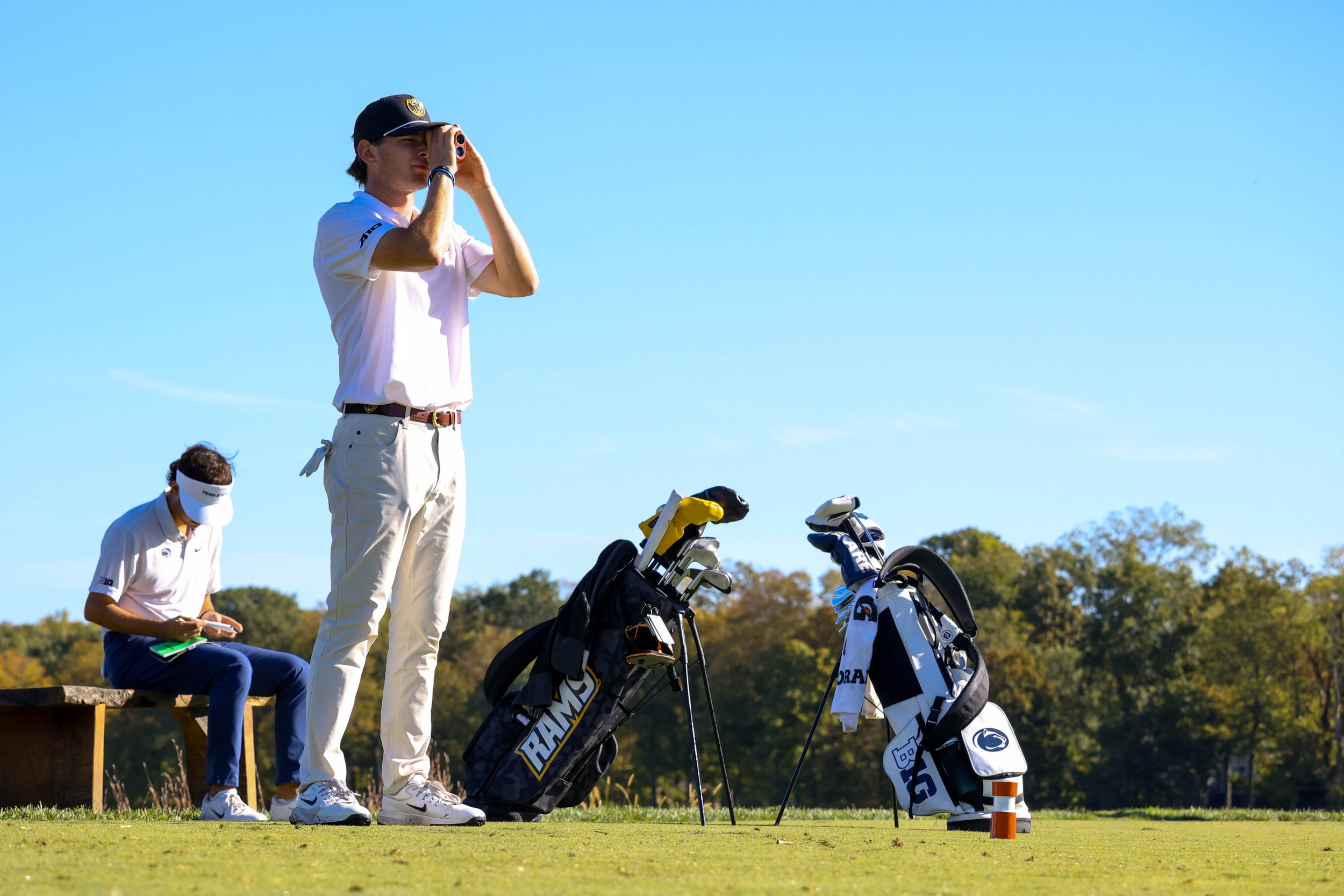 Two young male golfers on a golf course, one standing and using rangefinder and the other sitting and looking at a scorecard, with golf bags and clubs nearby on a clear, sunny day.