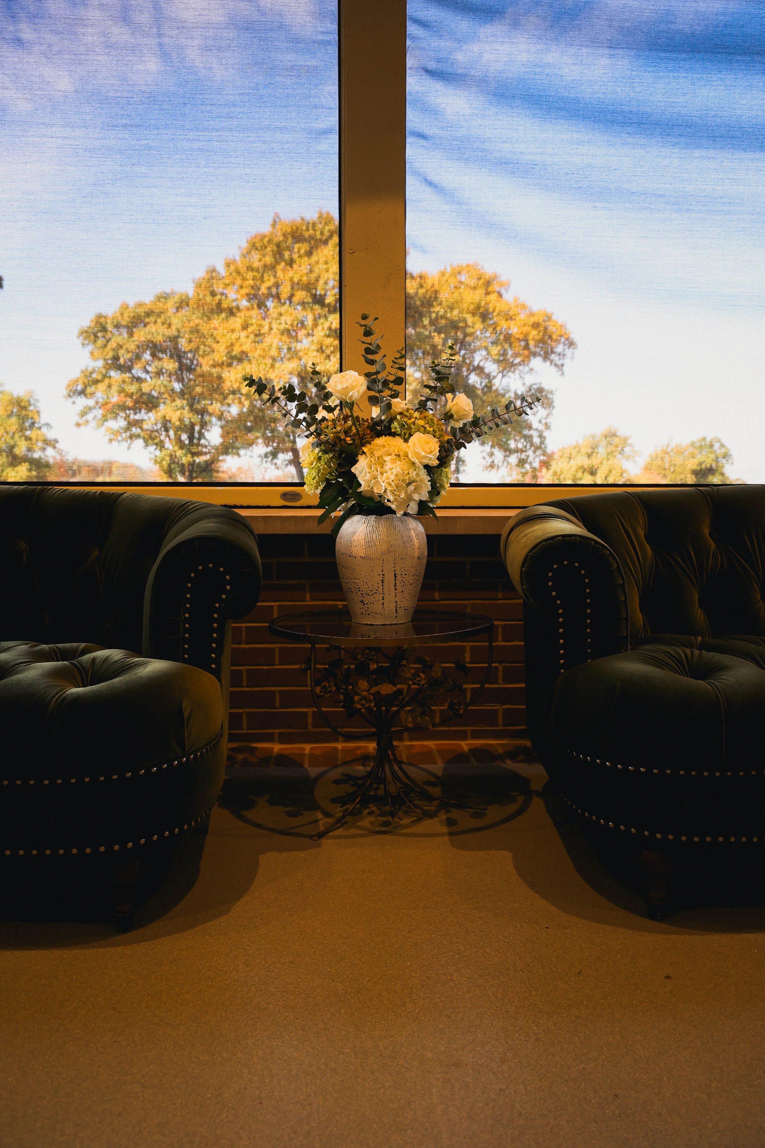 Two dark green leather tufted sofas facing each other with a small round table in between holding a white flower arrangement, placed in front of large windows showing trees and a blue sky.