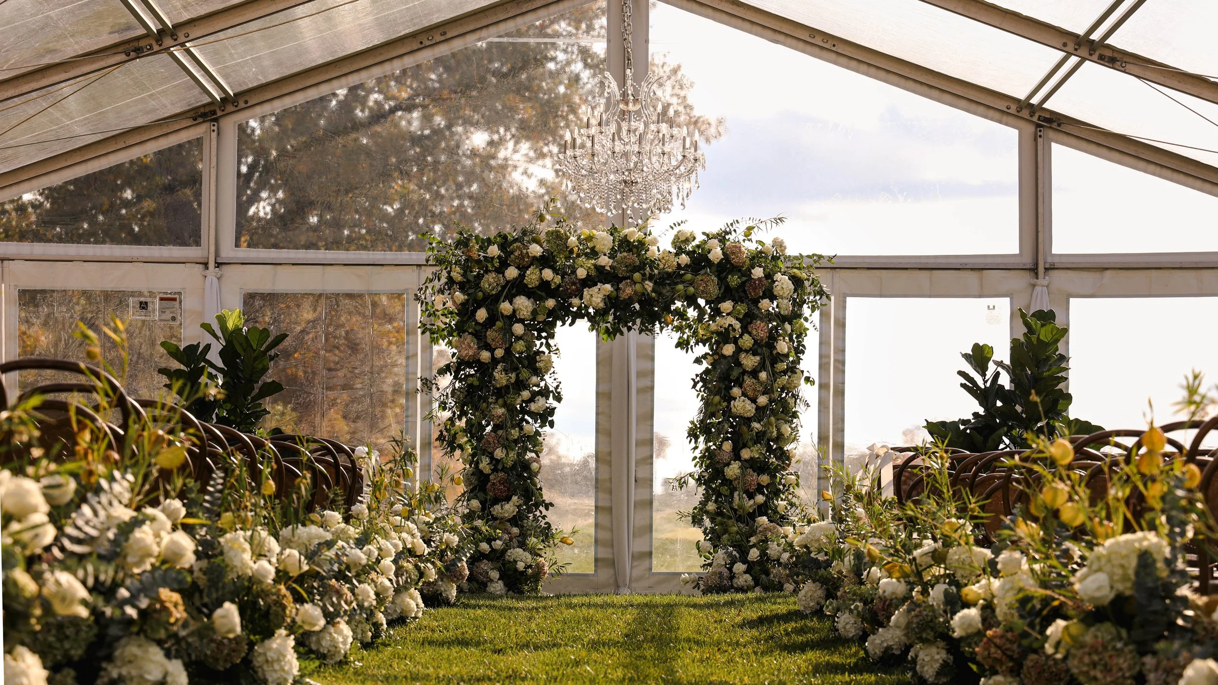 Flower arch and rows of white flowers inside a glass tent for a wedding ceremony.
