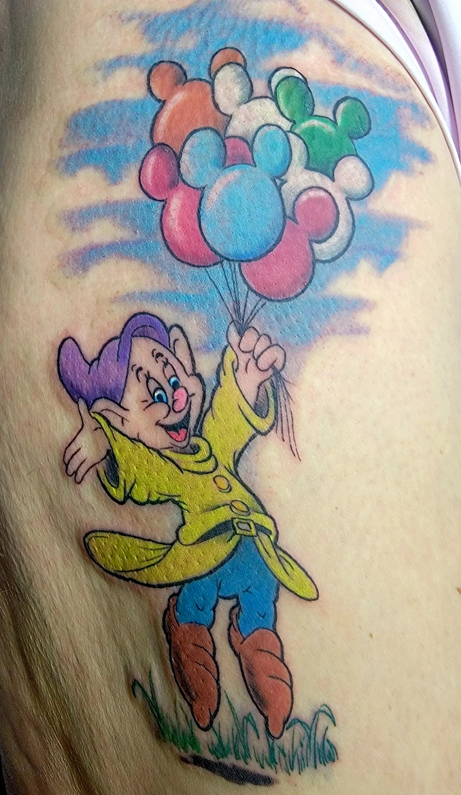 Colorful tattoo of a cartoon character, a cheerful boy with purple hair, yellow coat, blue pants, and brown boots, holding a bunch of multicolored balloons against a background of blue sky and clouds.