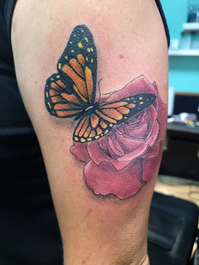 Tattoo of a monarch butterfly perched on a pink rose on a person's upper arm.