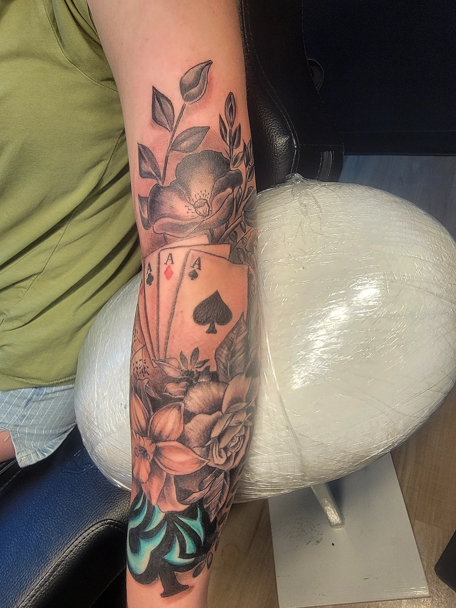 Tattoo of a large flower, a card with four aces, and leafy designs on a person's arm.