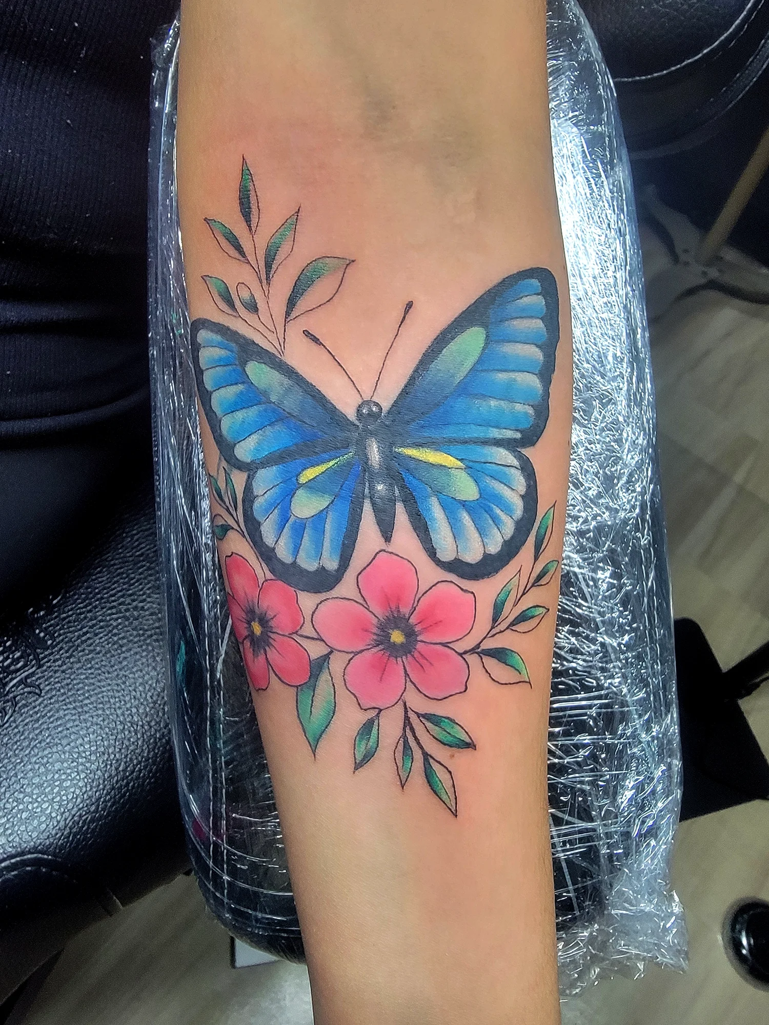 A colorful tattoo of a blue and yellow butterfly with pink flowers and green leaves on a person's forearm.