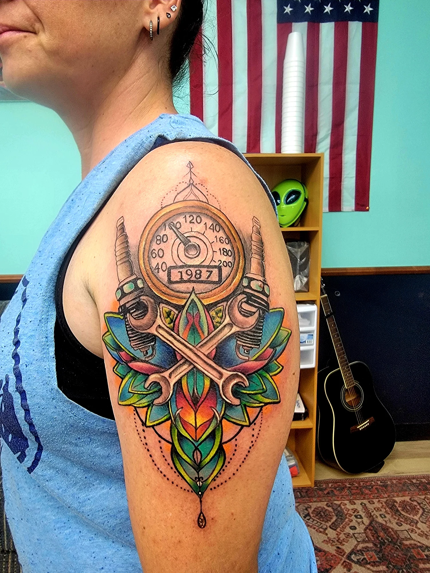 A colorful tattoo on a person's upper arm, featuring a pocket watch with the year '1987', crossed swords, winged elements, and a geometric pattern with bright colors. The person is wearing a blue sleeveless shirt, has multiple earrings, and has short