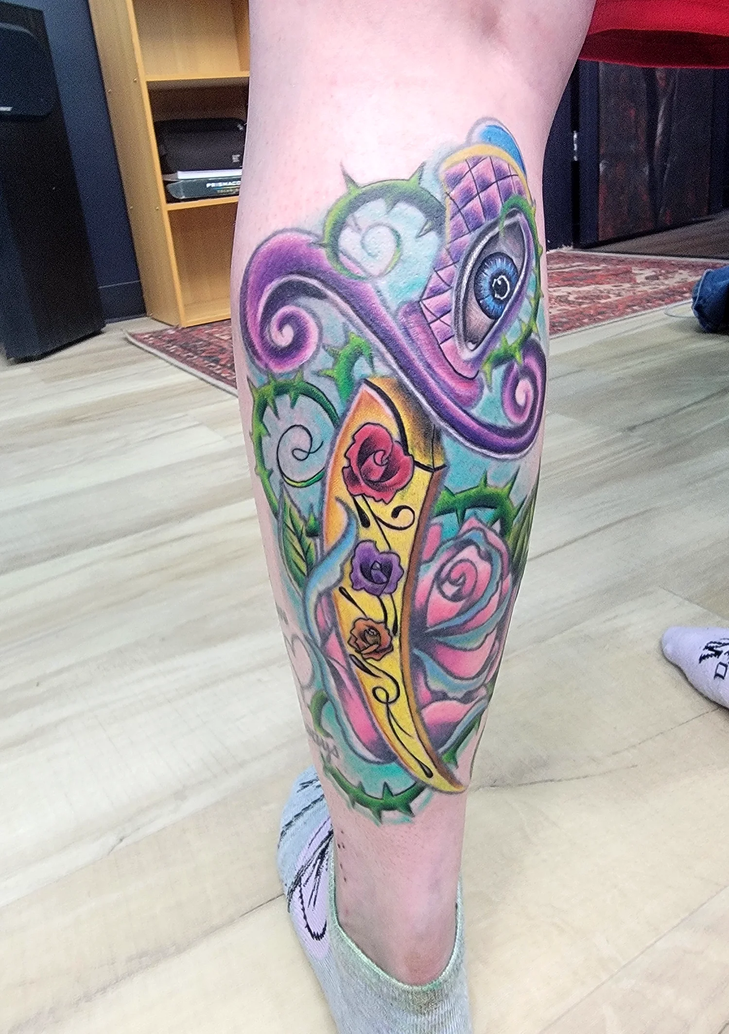 Colorful tattoo on a person's leg featuring a large eye, roses, and swirling patterns in purple, green, yellow, and pink.