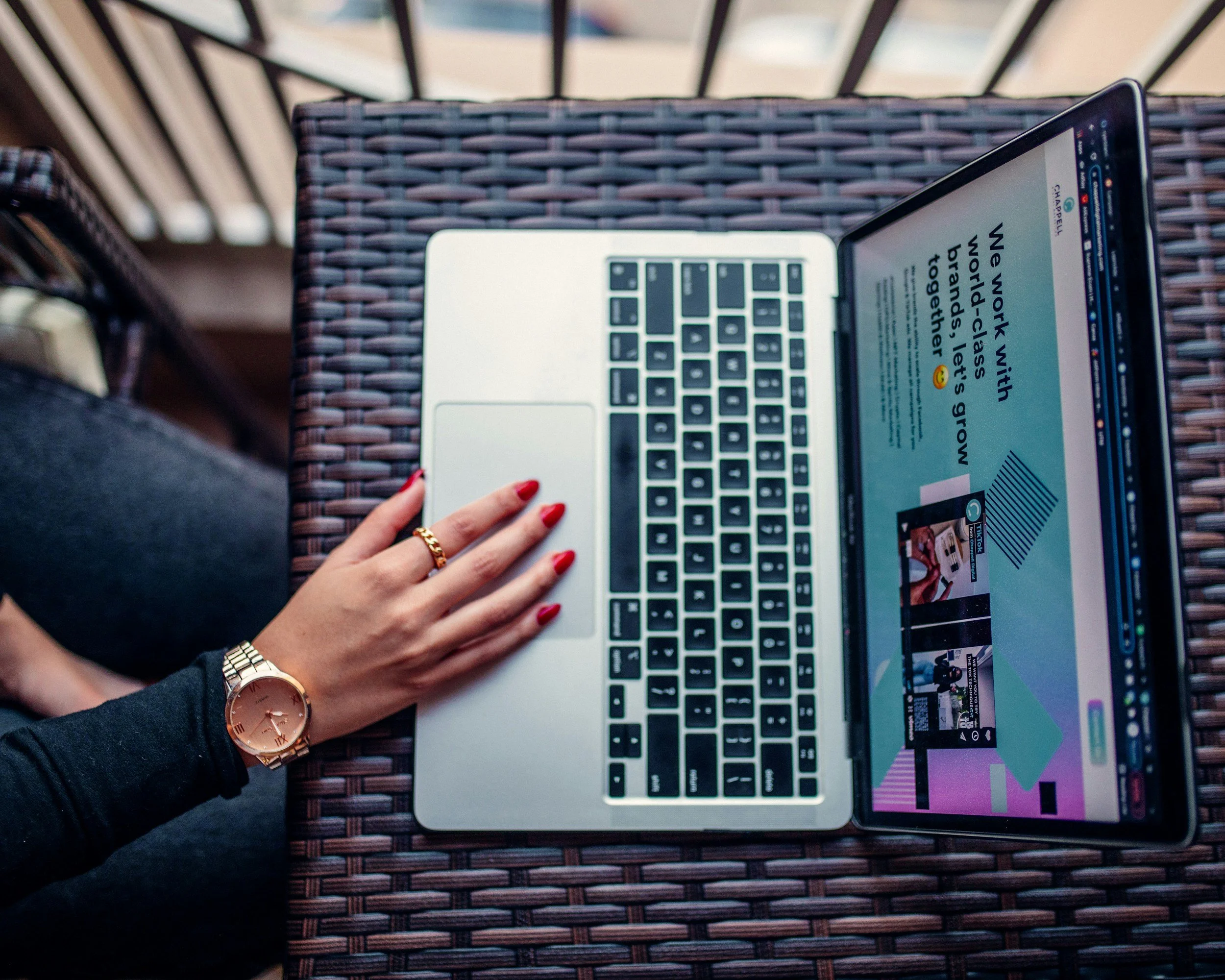 Persona con uñas rojas y reloj dorado usando una laptop en una mesa de mimbre, con pantalla mostrando una página web de estilo moderno y colorido.