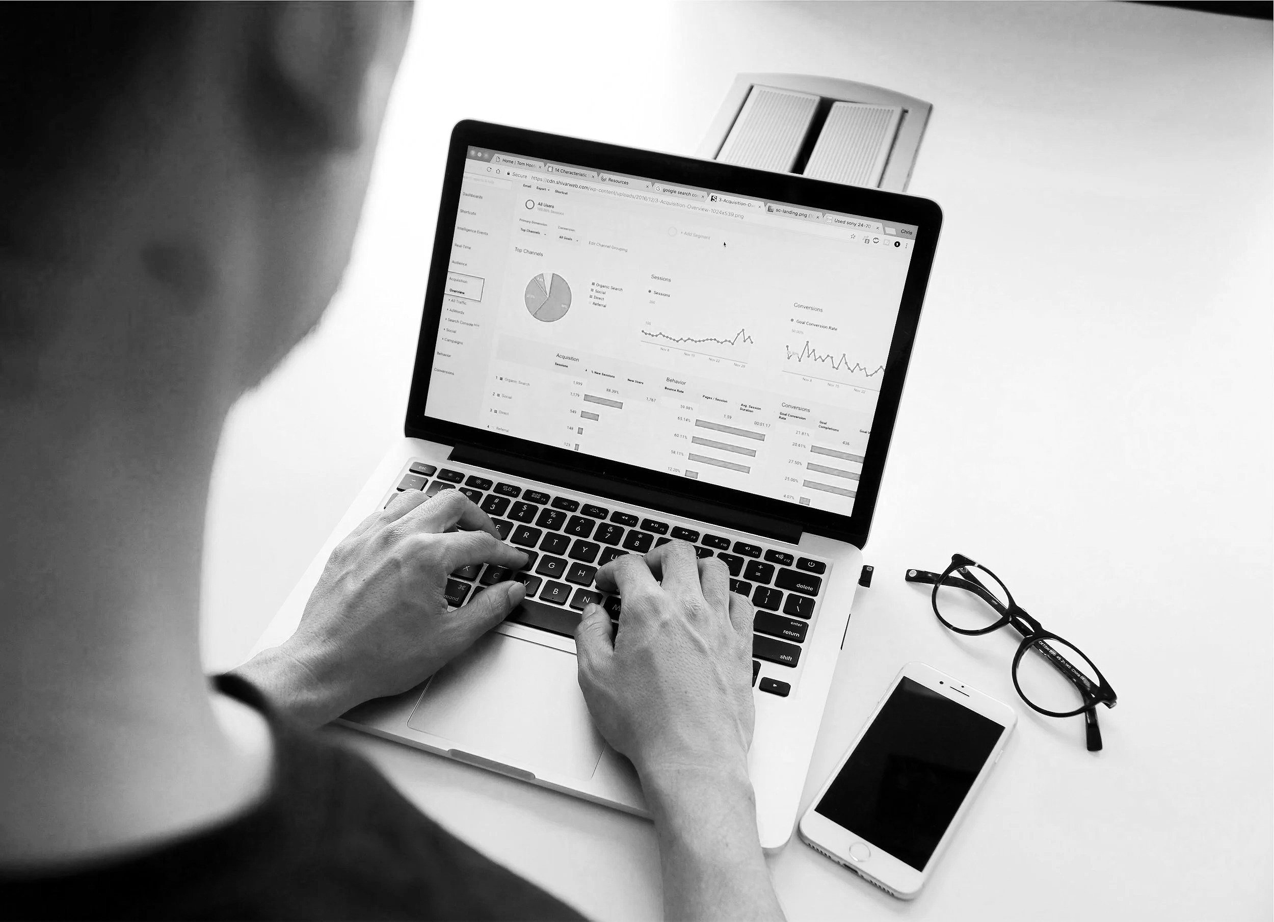 Persona trabajando en una computadora portátil con gráficos y datos en pantalla, en una mesa blanca, acompañada de gafas y teléfono móvil.