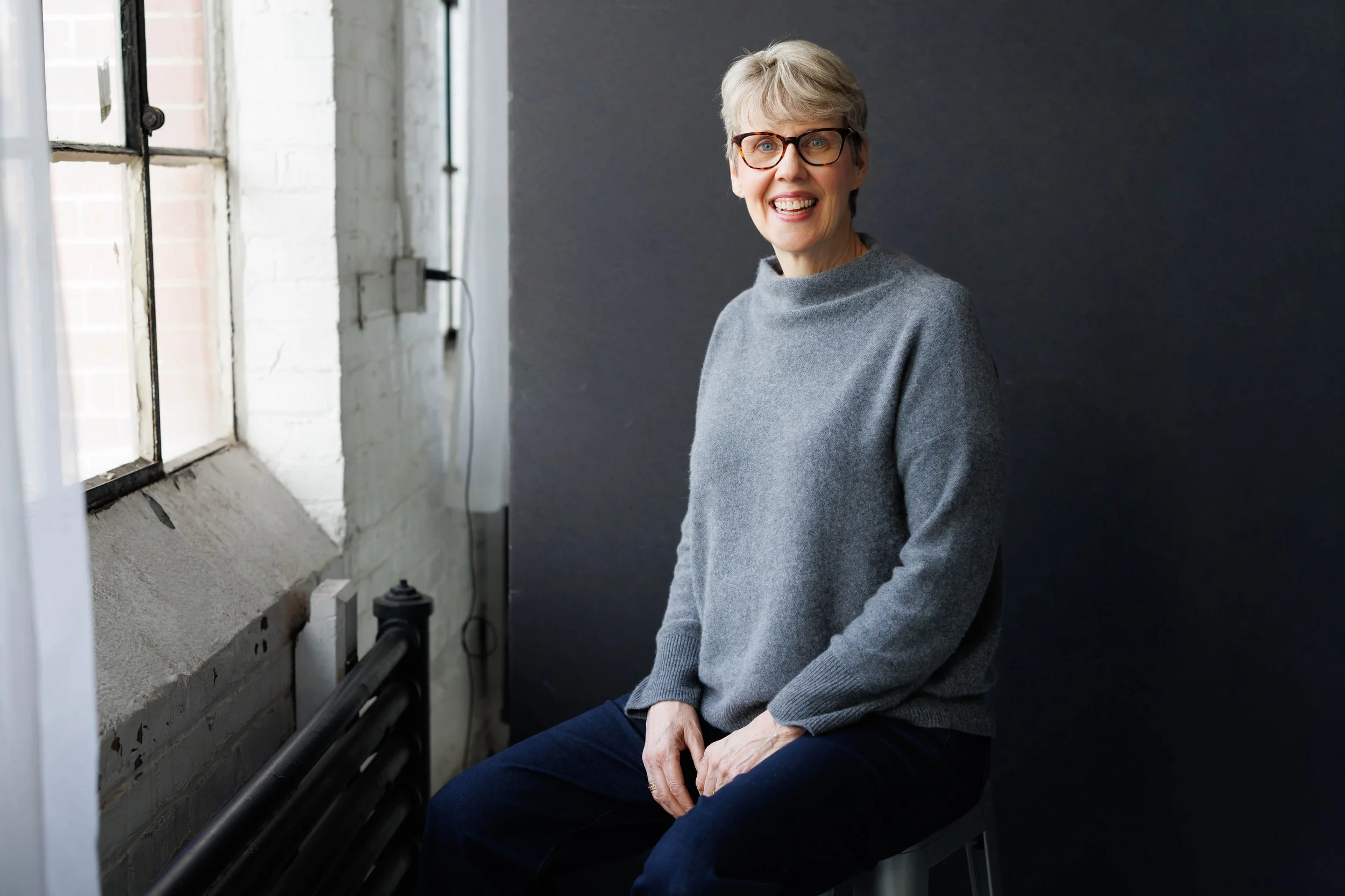 An elderly woman sitting by a window in a room with a dark wall, smiling and wearing glasses and a gray sweater.